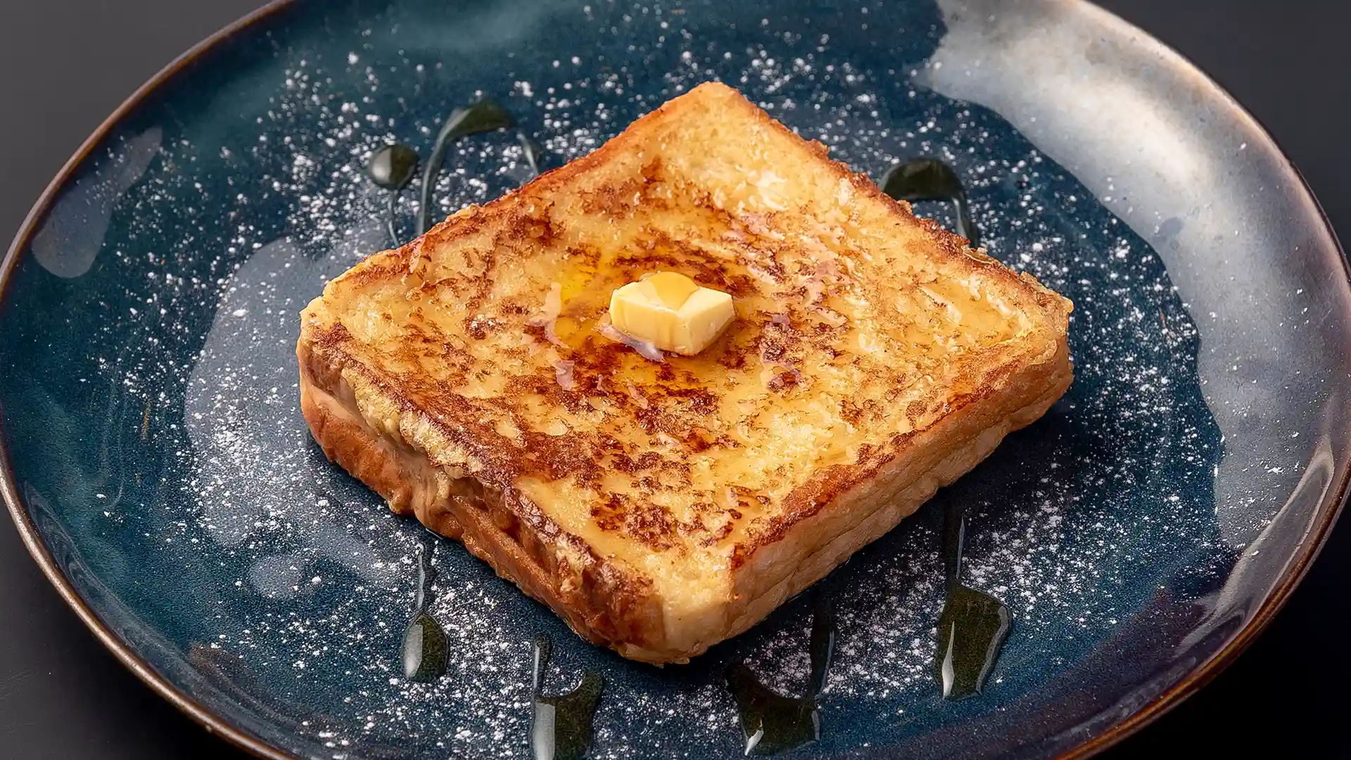 Hong Kong French toast on a blue plate, drizzled with golden syrup, dusted with icing sugar, and topped with a knob of butter.