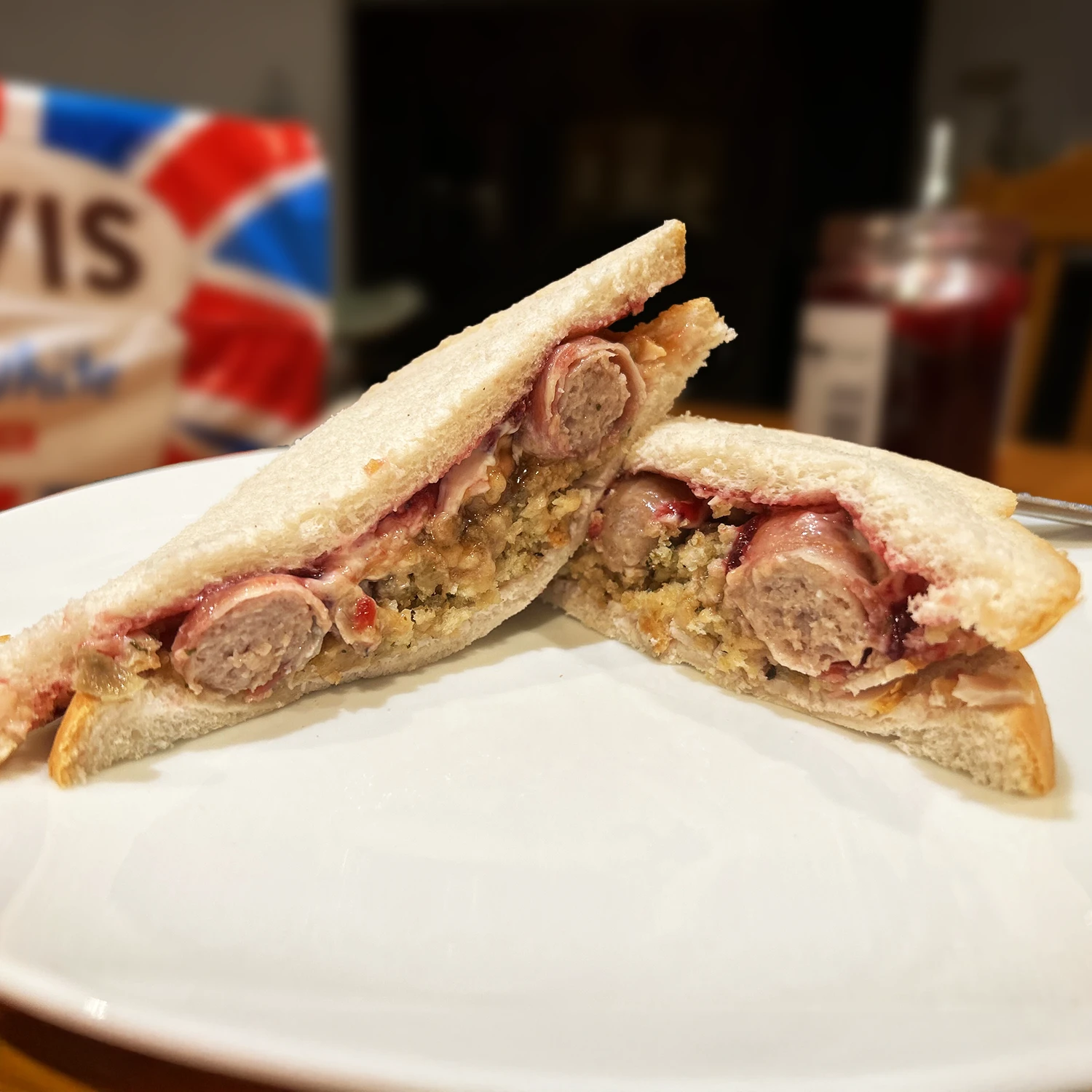 A Hovis Soft White Medium Sliced sandwich sliced in half on a white plate, filled with turkey slices, stuffing, cranberry sauce and pigs in blankets.