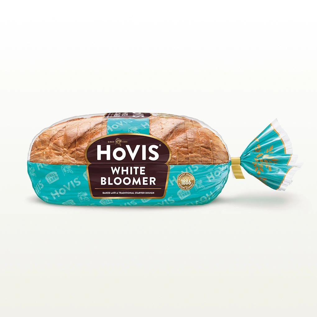 Hovis White Bloomer bread on a white backdrop with a subtle shadow. The packaging is half transparent and half light teal, decorated with golden wheats.