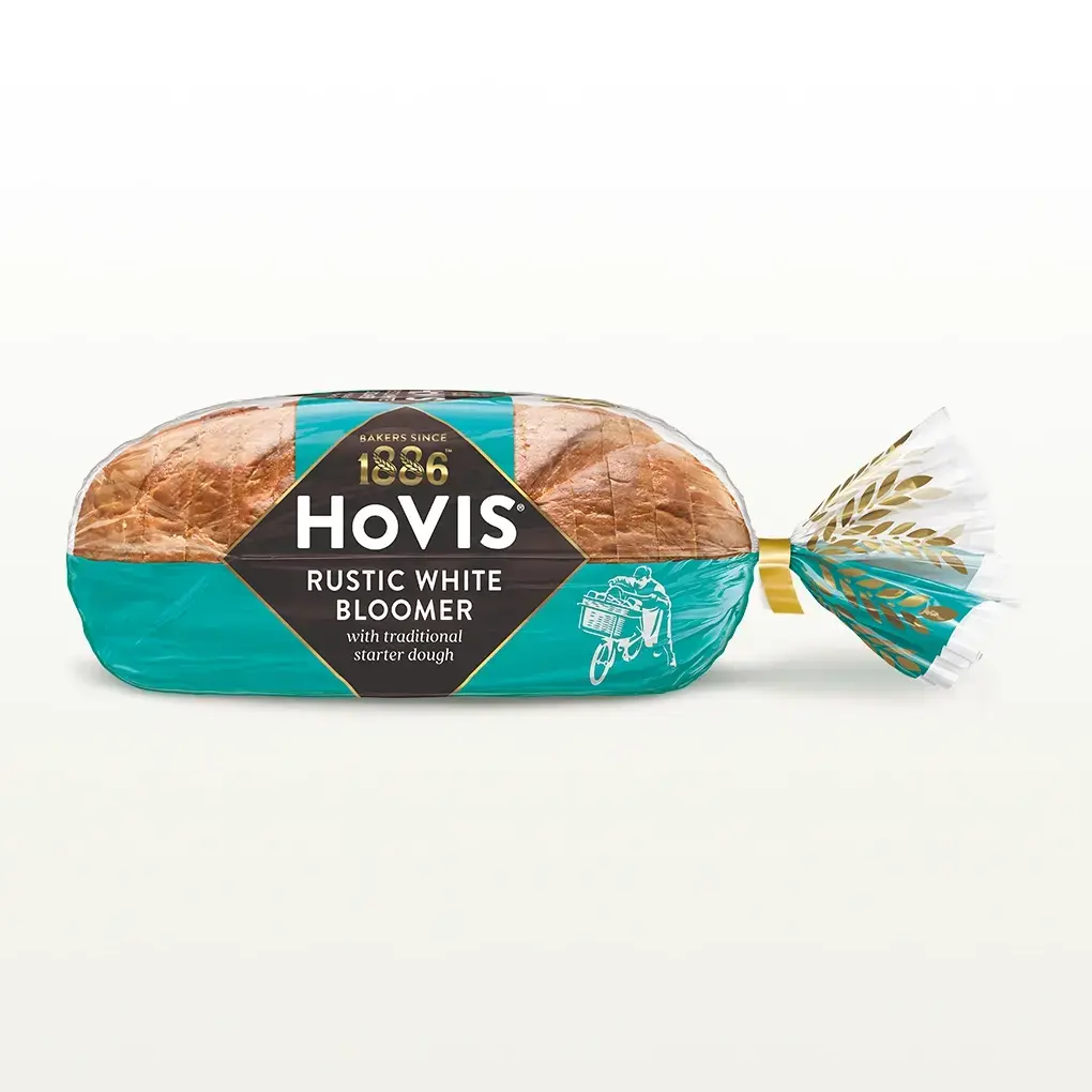 Hovis White Bloomer bread on a white backdrop with a subtle shadow. The packaging is half transparent and half light teal, decorated with golden wheats.