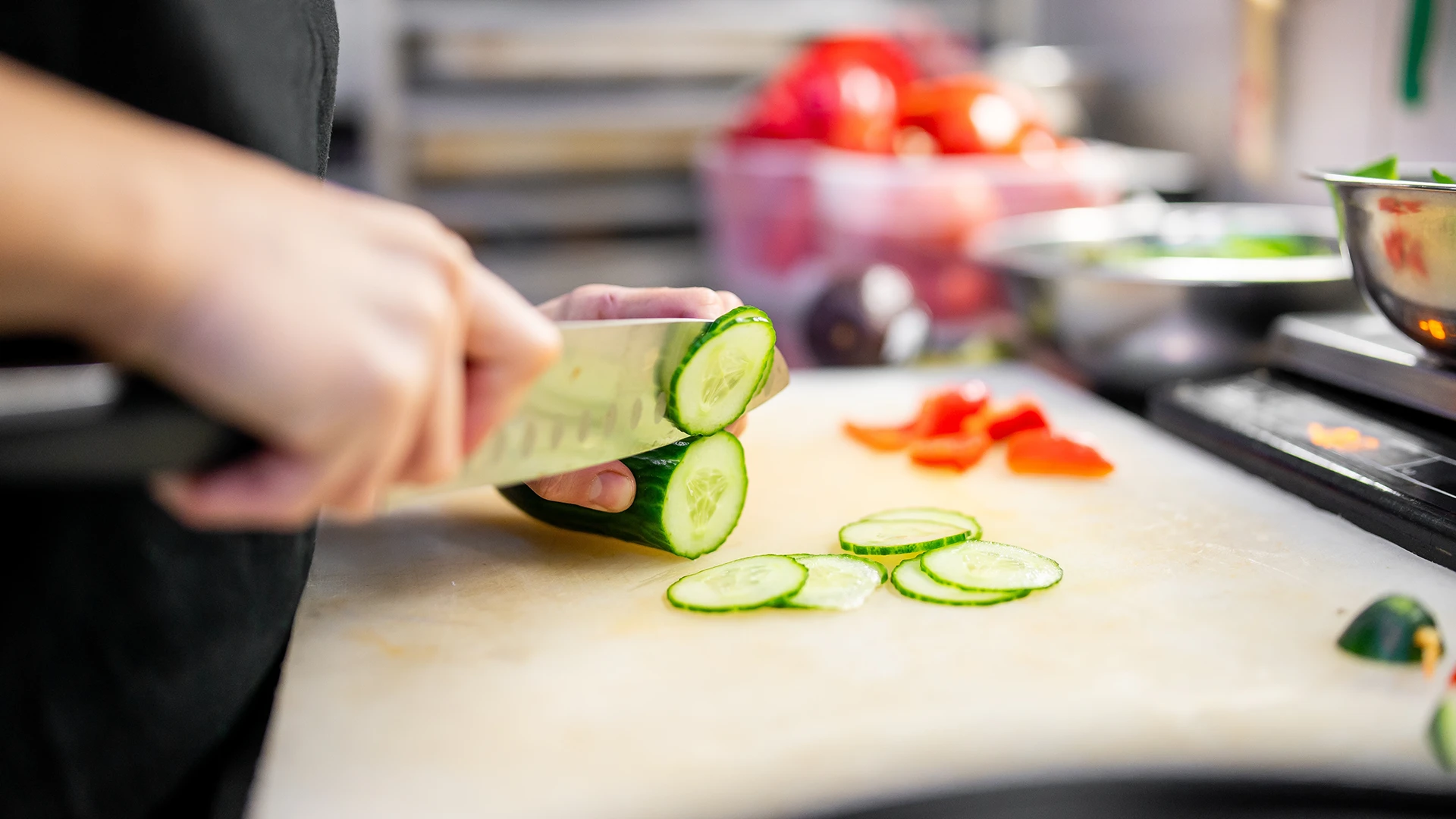 Hands using a sharp knife to thinly slice a cucumber on a wooden chopping board.