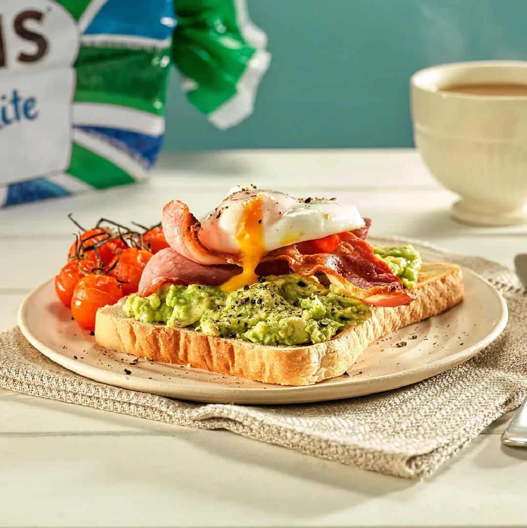 A slice of white bread on a plate, topped with mashed avocado, crispy bacon, a poached egg with yellow yolk running and some grilled vine cherry tomatoes on the side. 