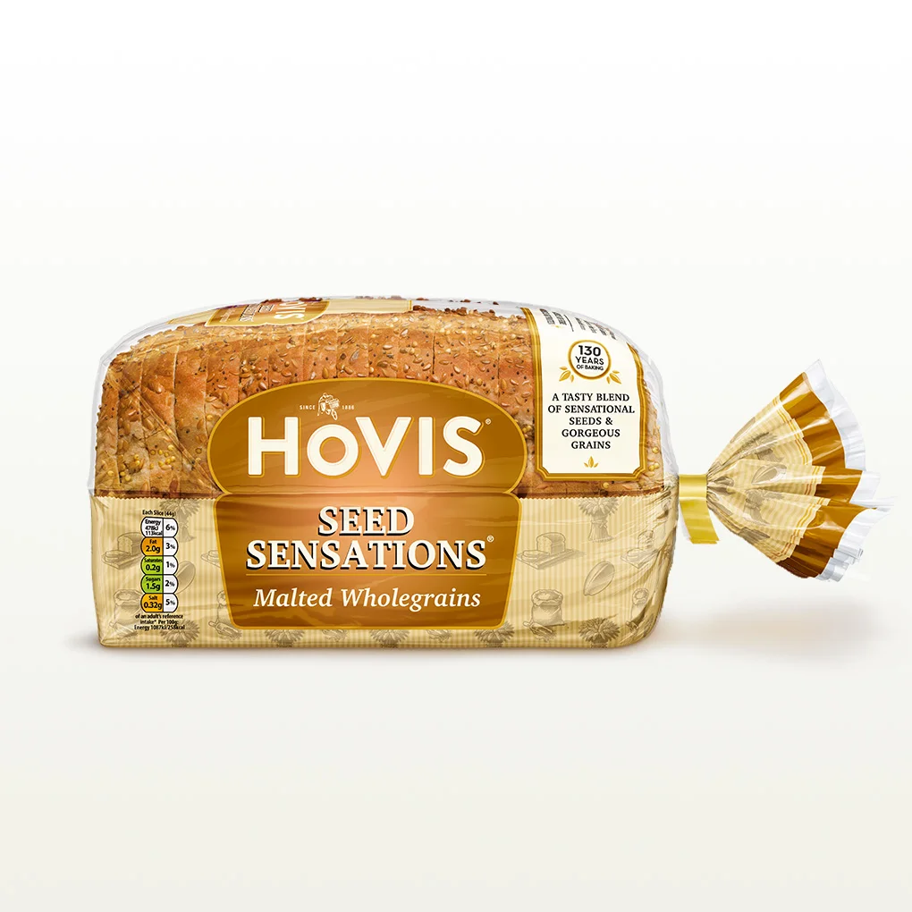 Hovis Seed Sensations Malted Wholegrain bread on a white backdrop with a subtle shadow. The packaging is half transparent and half beige, decorated with images of sliced bread, seeds and wheat.