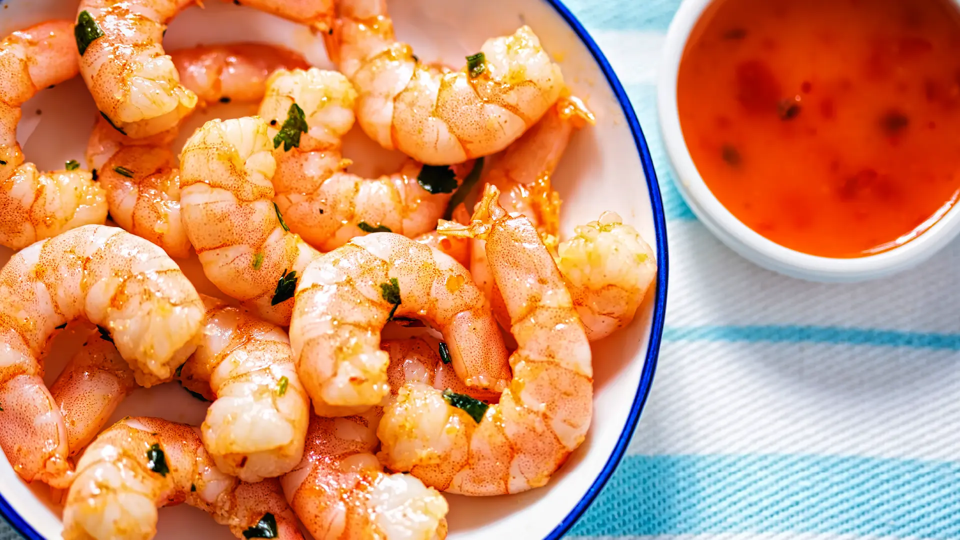 A top view of a bowl of prawns topped with chopped greens, with a small bowl of chilli sauce on the side.