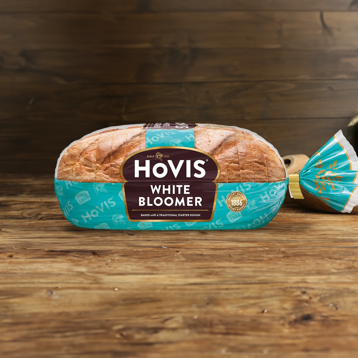 Hovis White Bloomer on a wooden table, with a bowl of wheat grains, a wooden scoop, and dried wheat in the background.