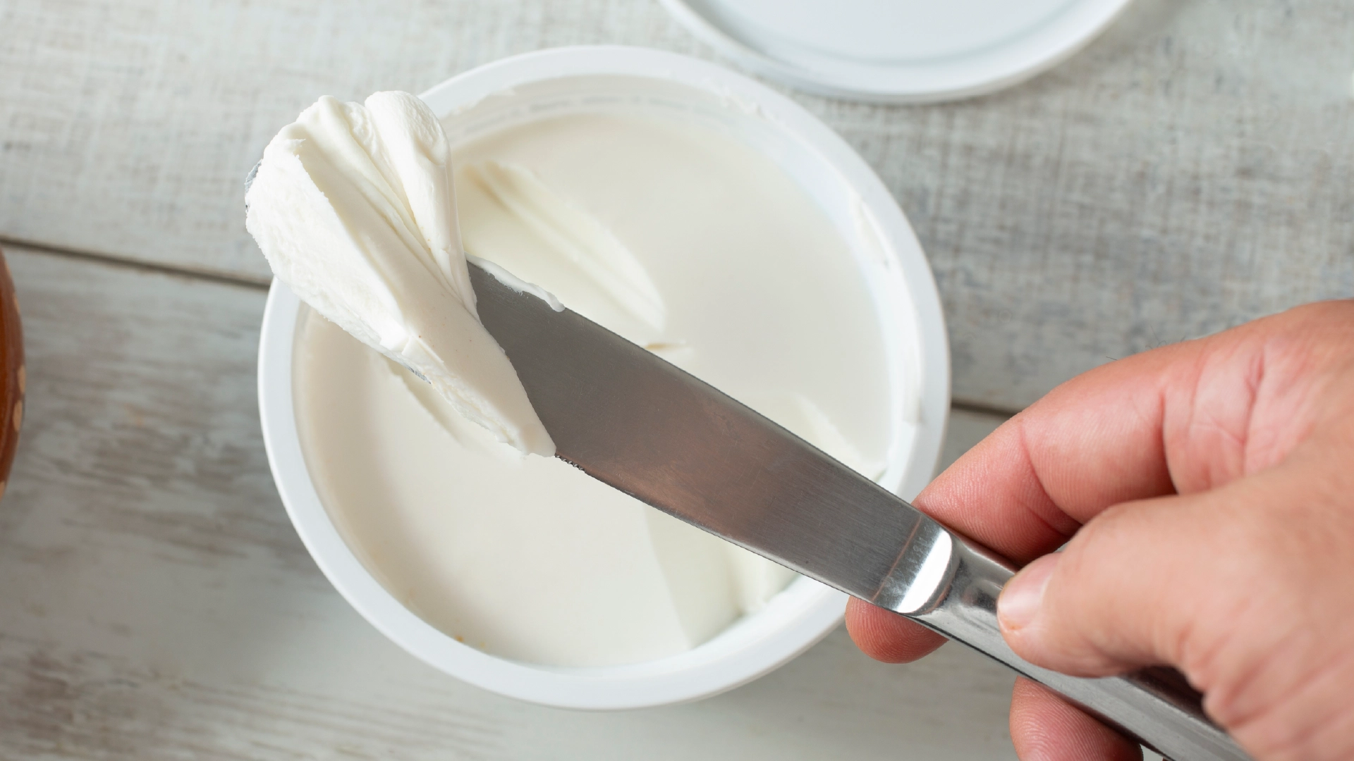 A hand holding a knife to scoop white cream cheese from a pot.