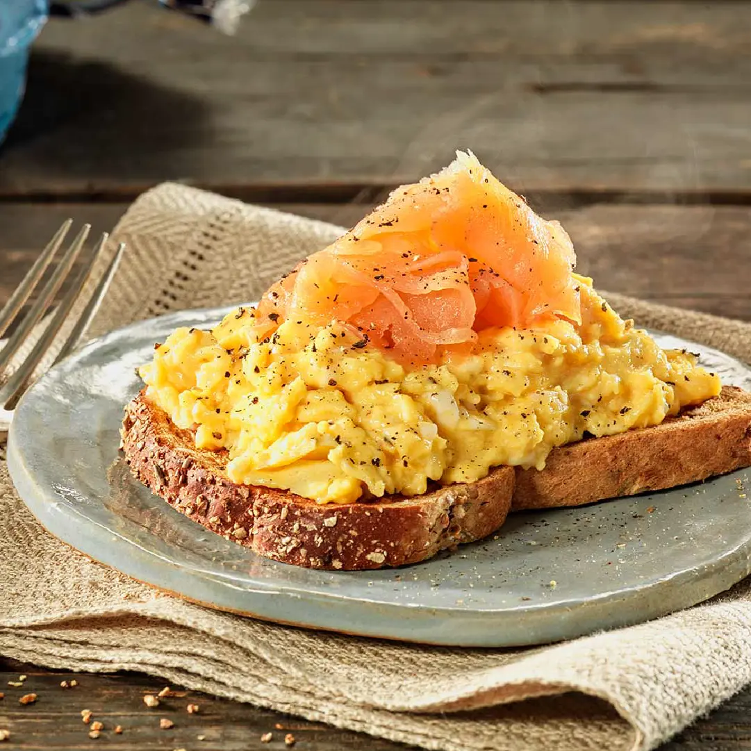 A slice of brown bread on a plate, topped with scrambled eggs, slices of smoked salmon, and a sprinkle of black pepper.