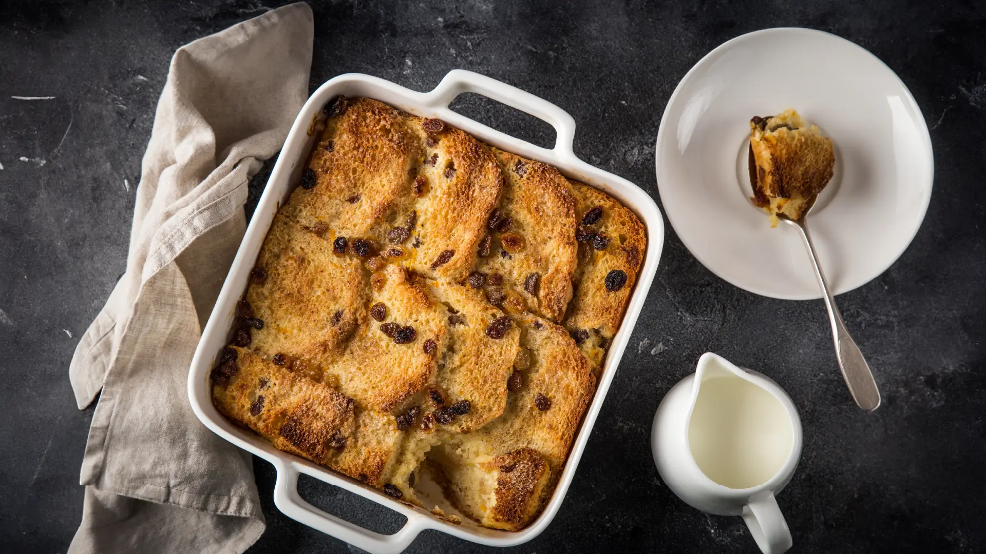 A top-down view of a square baking tray with layered toasted bread topped with raisins. Next to it, a small plate with a spoonful of bread pudding.