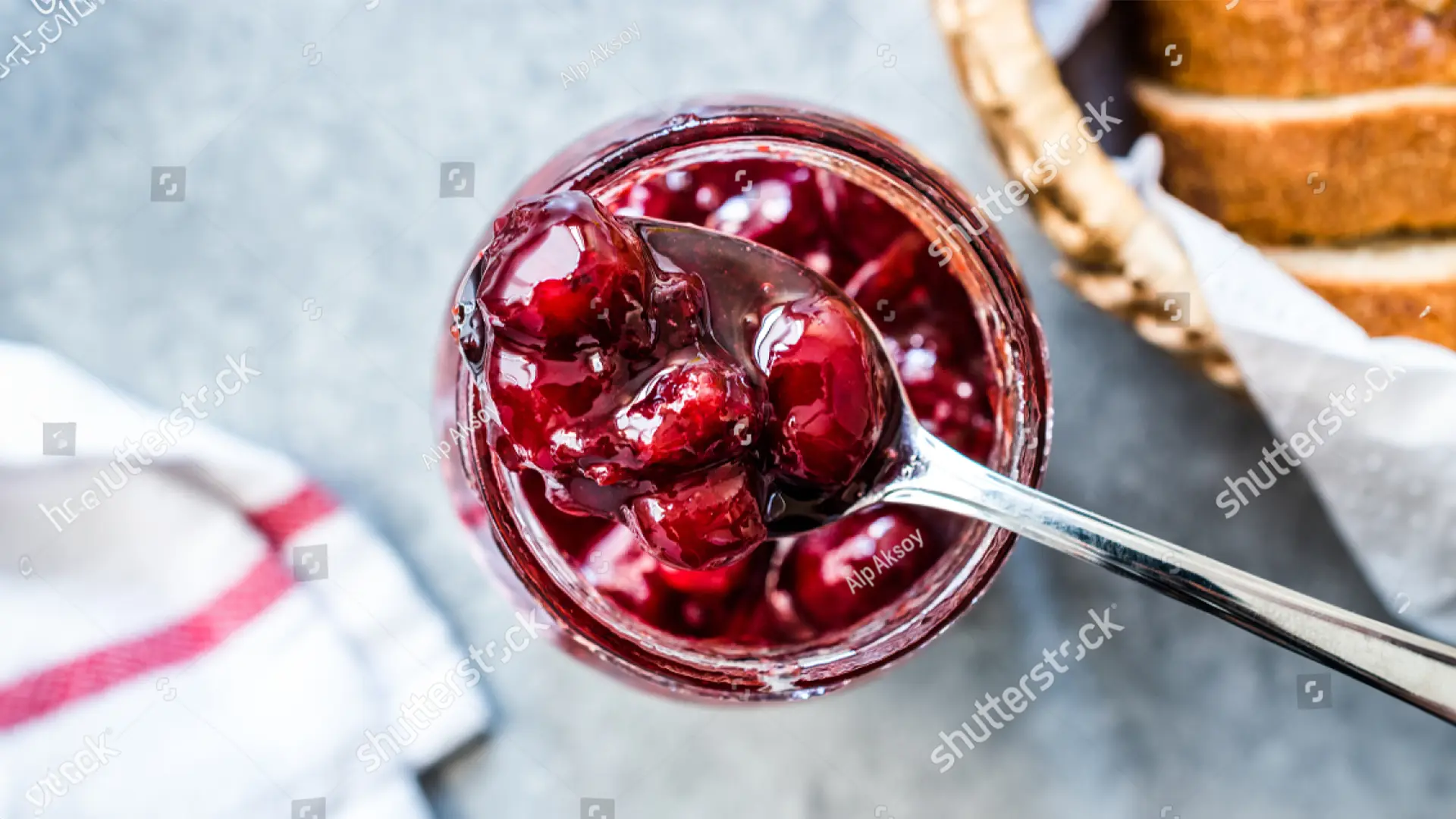 A top-down view of a small glass jar filled with cranberry sauce and a metal spoon scooping some of the sauce from it. In the background, some slices of brown bread rest in a wooden basket.