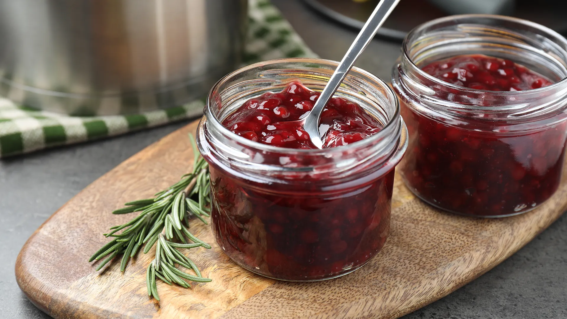 A top-down view of a small glass jar filled with cranberry sauce and a metal spoon scooping some of the sauce from it. In the background, some slices of brown bread rest in a wooden basket.