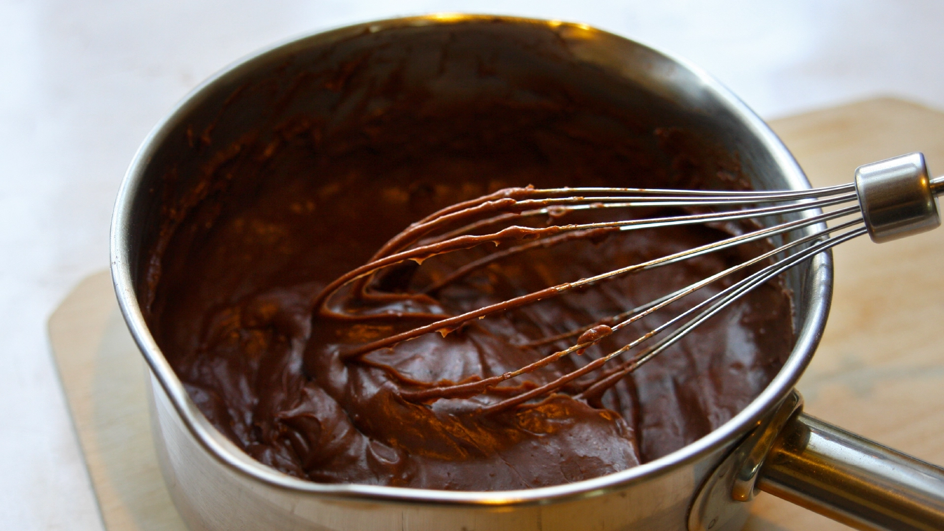 A medium pot filled with molten brown chocolate and a metal whisk inside.