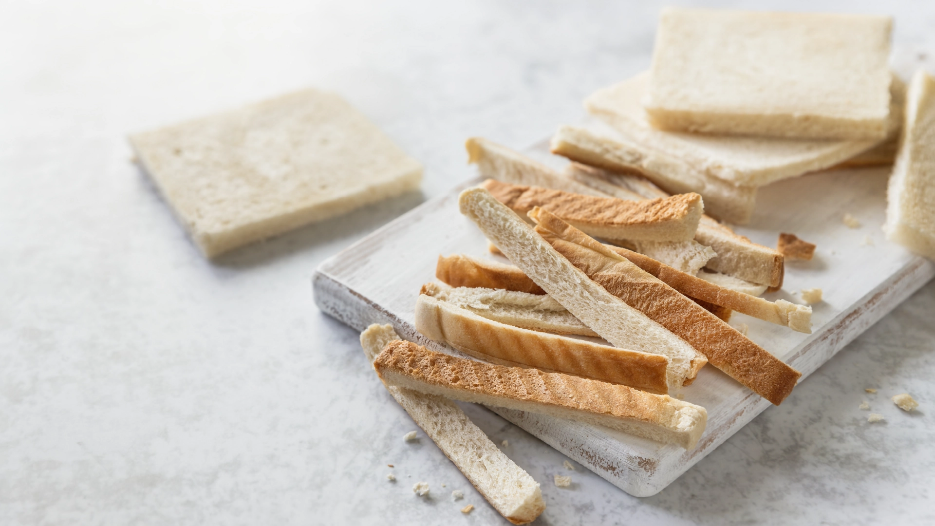 Slices of white bread with the crusts removed, placed on a cutting board with the crusts set aside.