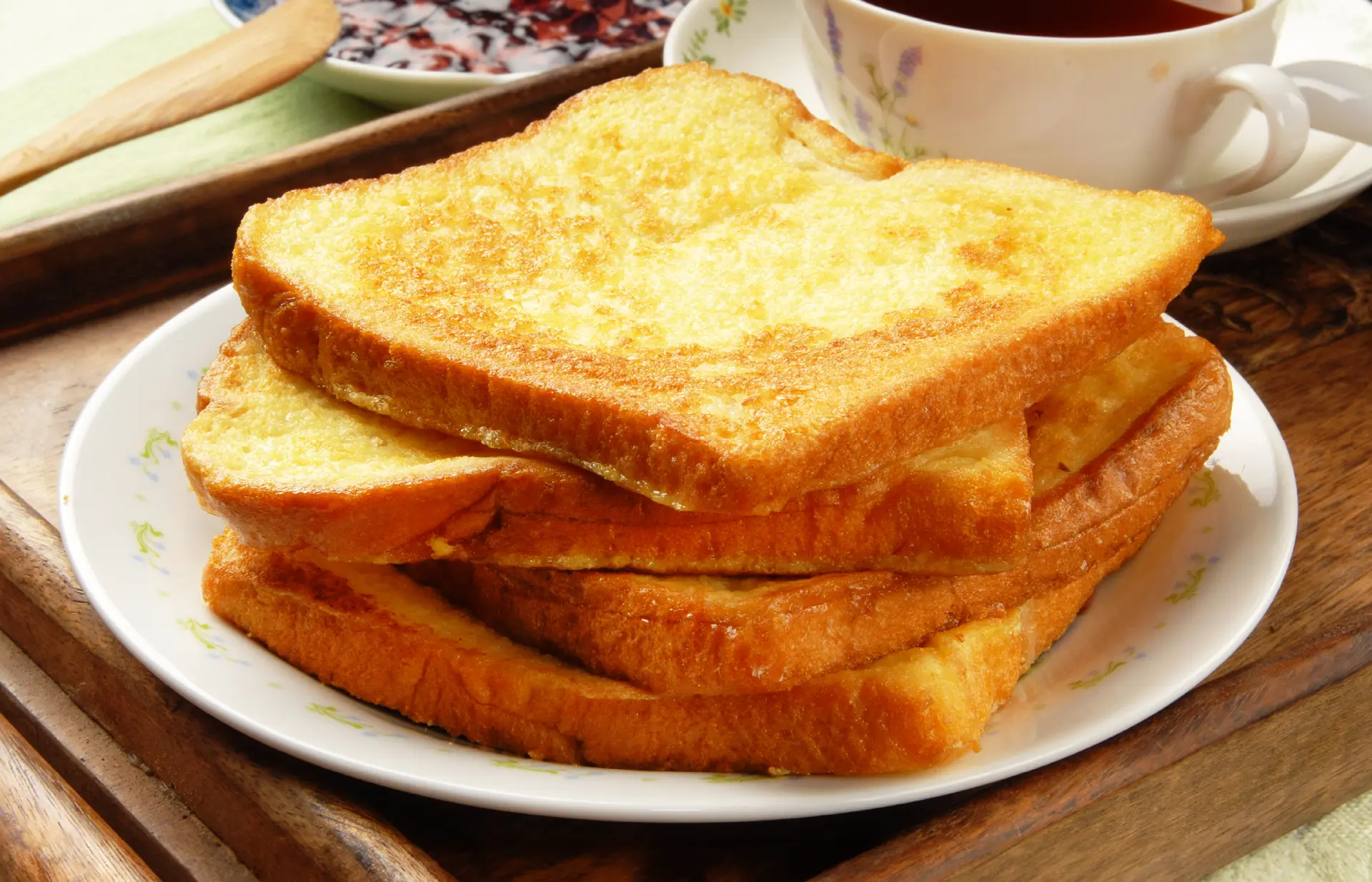 Four slices of toasted white bread stacked on a plate, with a cup of tea and a small plate of jam on the side.