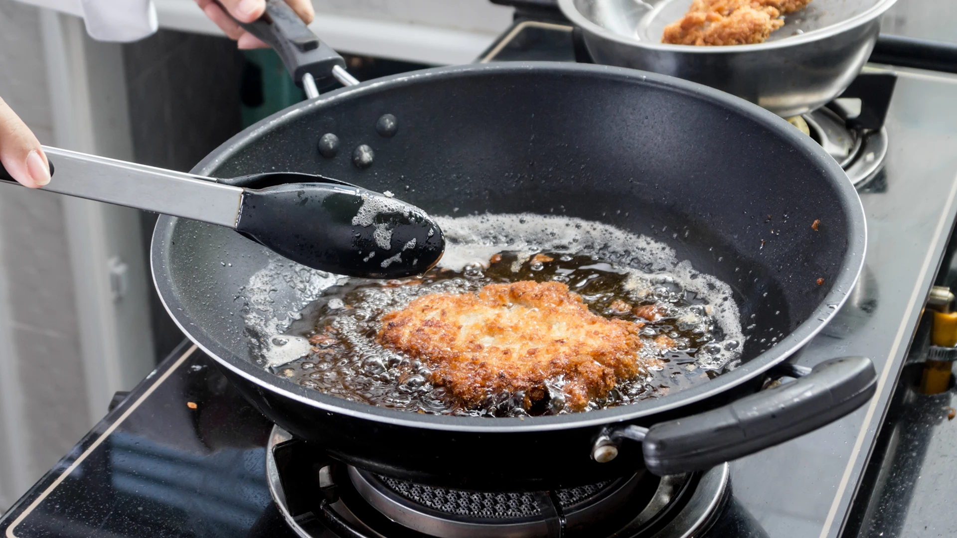 A hand holds the handle of a frying pan, deep frying slightly browned, crumb-coated chicken, while the other hand holds kitchen tongs.