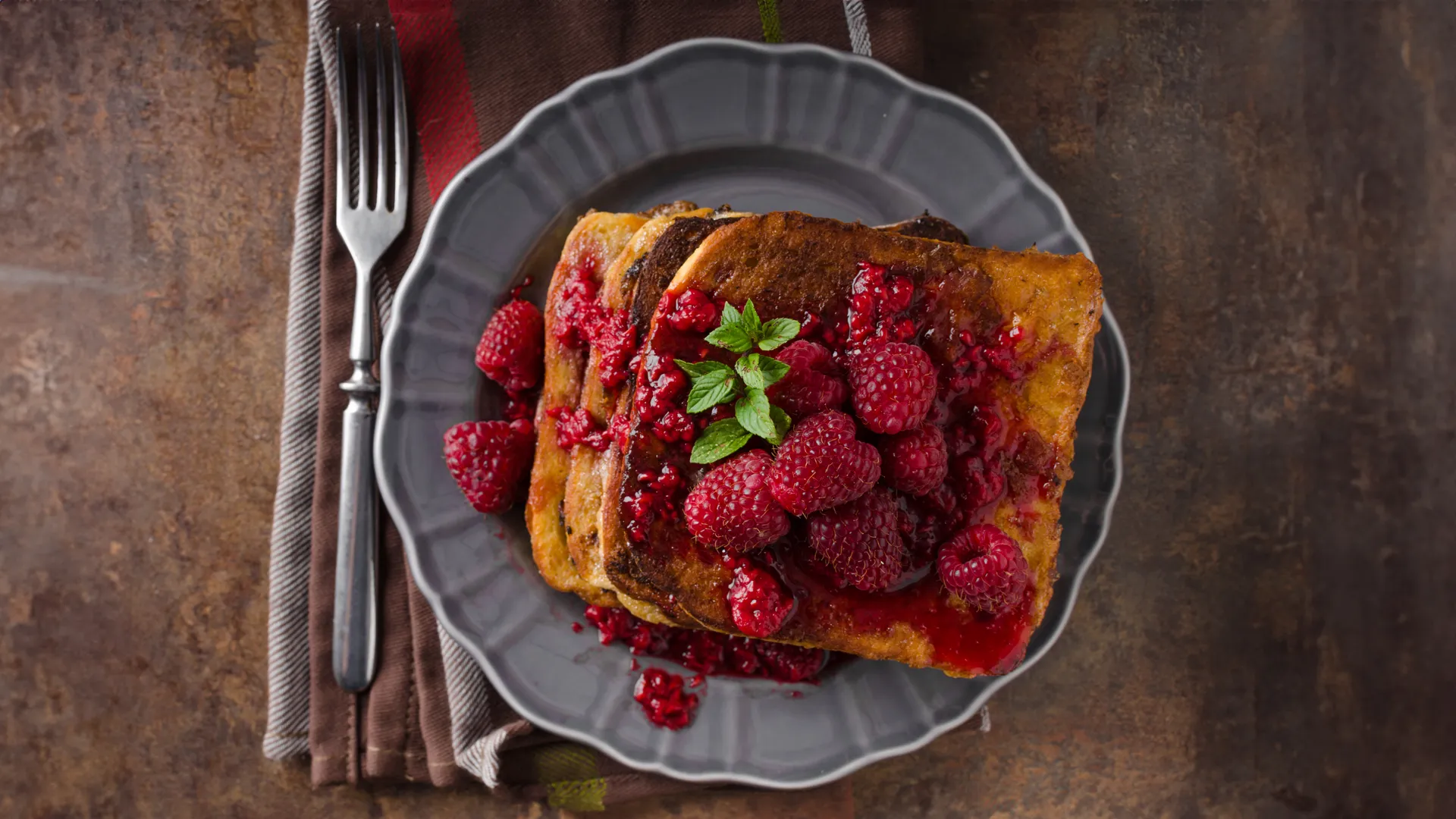 A grey plate on a dark brown table, with four golden Hovis Soft White Medium Sliced French toasts stacked. Some raspberries and a mint leaf on top.