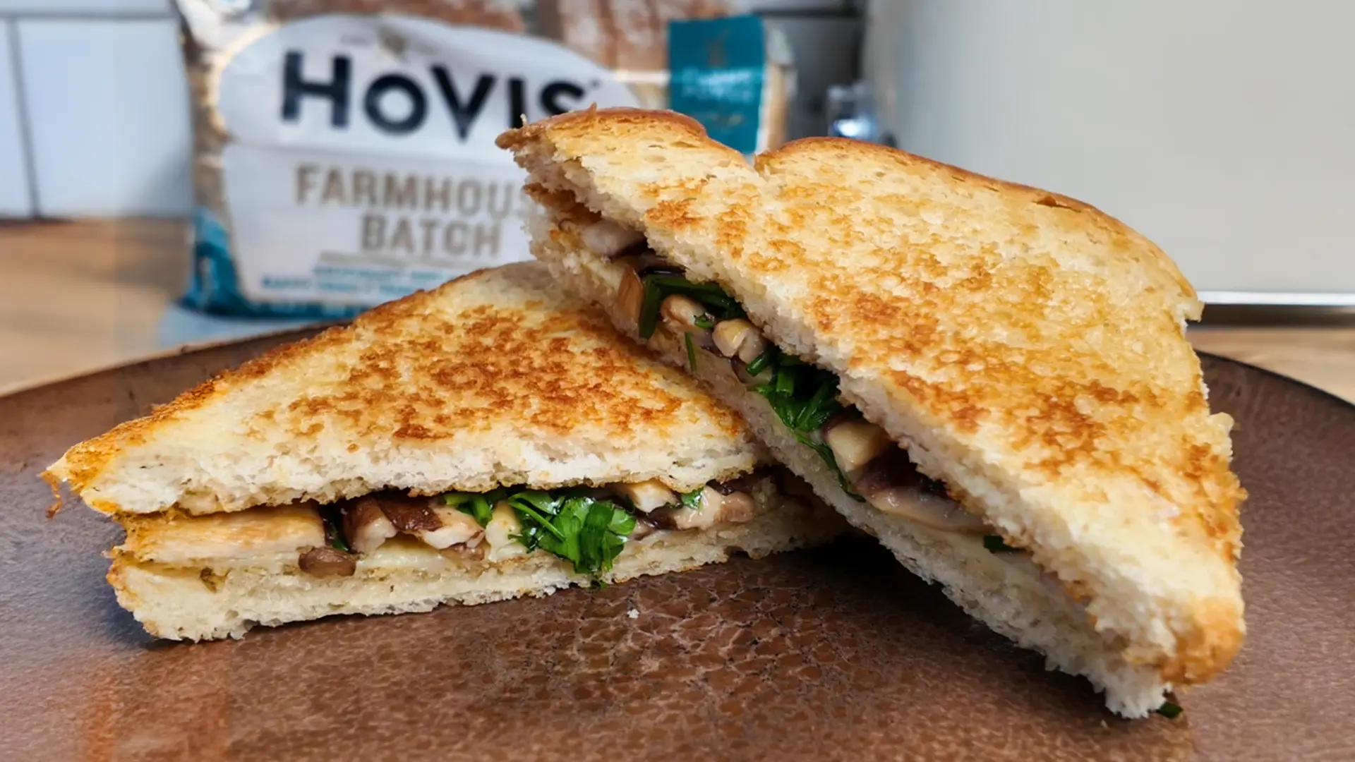 A golden-brown toasted sandwich cut in half, revealing a filling of sautéed mushrooms, greens, and melted cheese. In the background, a Hovis Farmhouse Batch bread pack is visible on a kitchen counter.