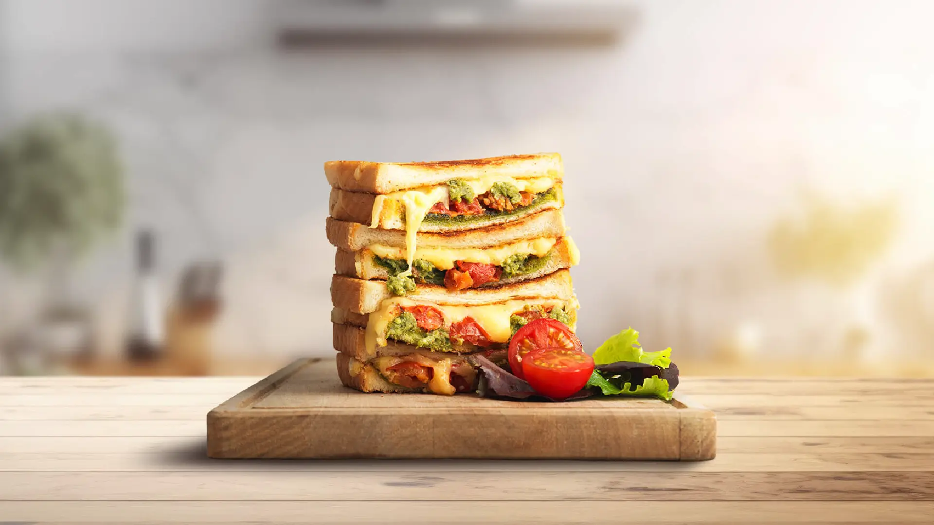 Four halves of pesto and cheese toasties stacked on a wooden chopping board, with molten cheese and pesto slightly visible between the slices. A halved tomato and some salad leaves are placed to the right.