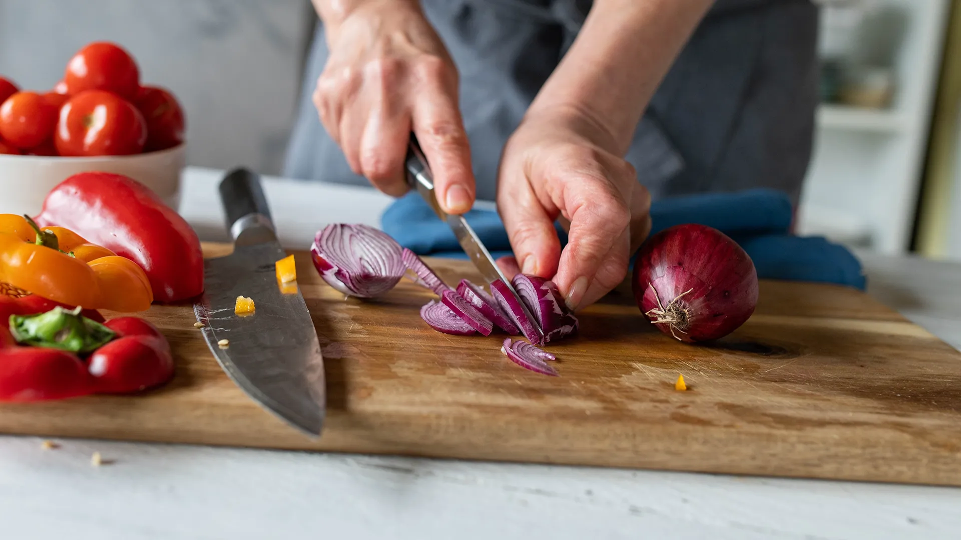 Someone uses a sharp knife to cut a red onion into thin slices on a wooden chopping board, with a bowl of tomatoes and yellow and red bell peppers beside them.