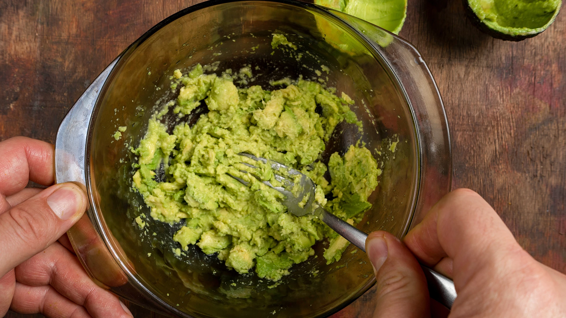 A hand holds a glass bowl of mashed avocado while the other hand uses a fork to mash it.