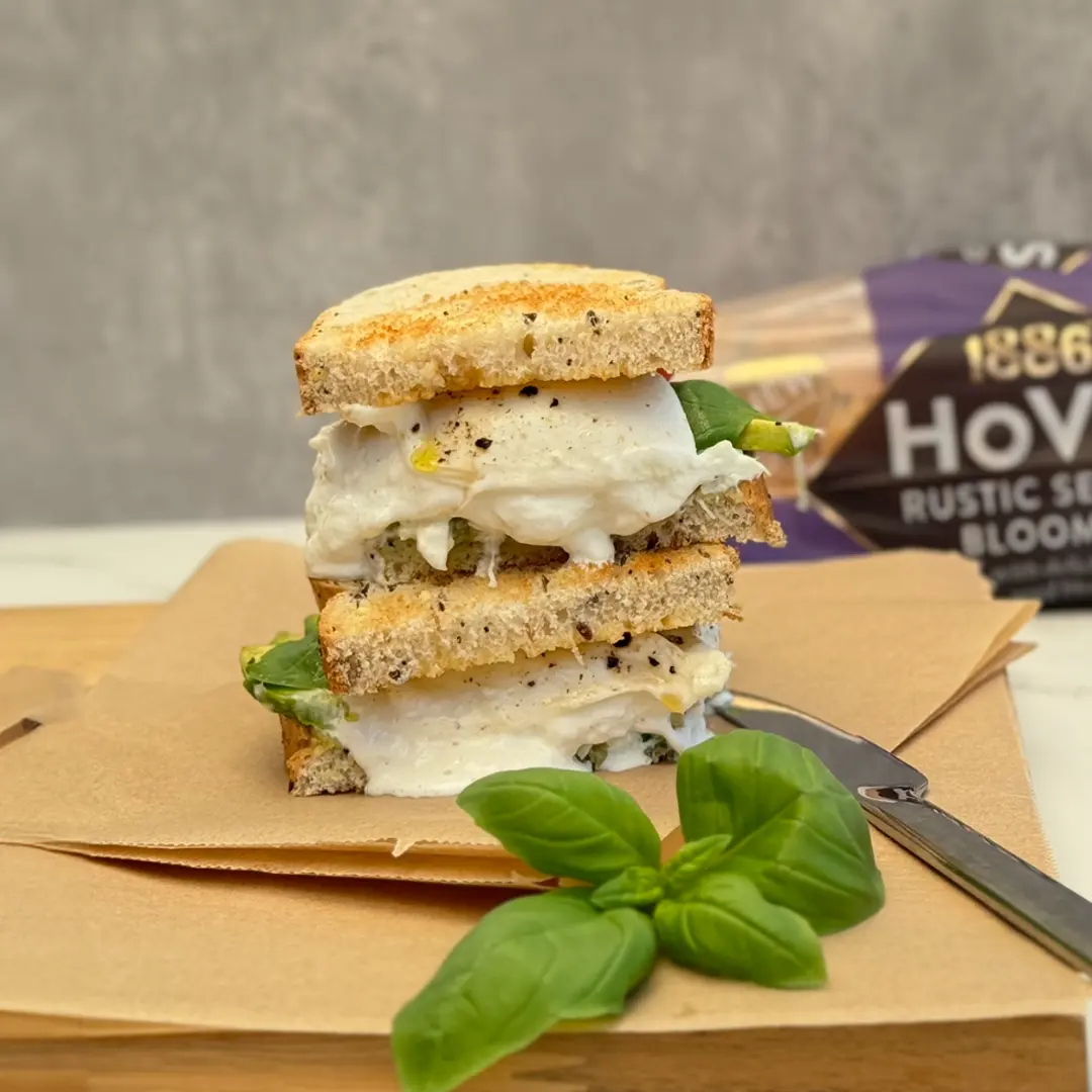 Two halves of a toasted sandwich with creamy white cheese, basil leaves, avocado and small pieces of ground black pepper oozing out. In the foreground, additional basil leaves and a knife are visible.