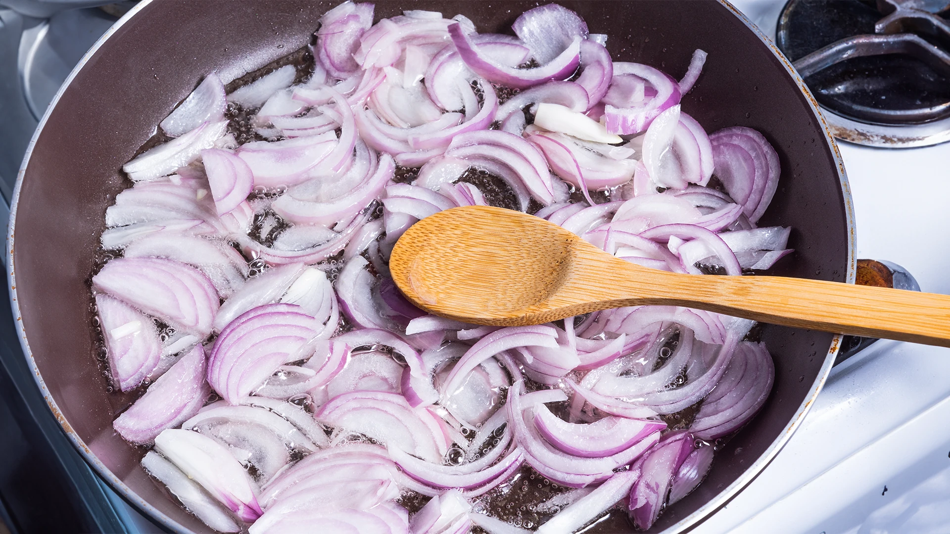 A top-down view of someone frying chopped red onions in a pan with oil on the hob, with a wooden spoon resting inside the pan.