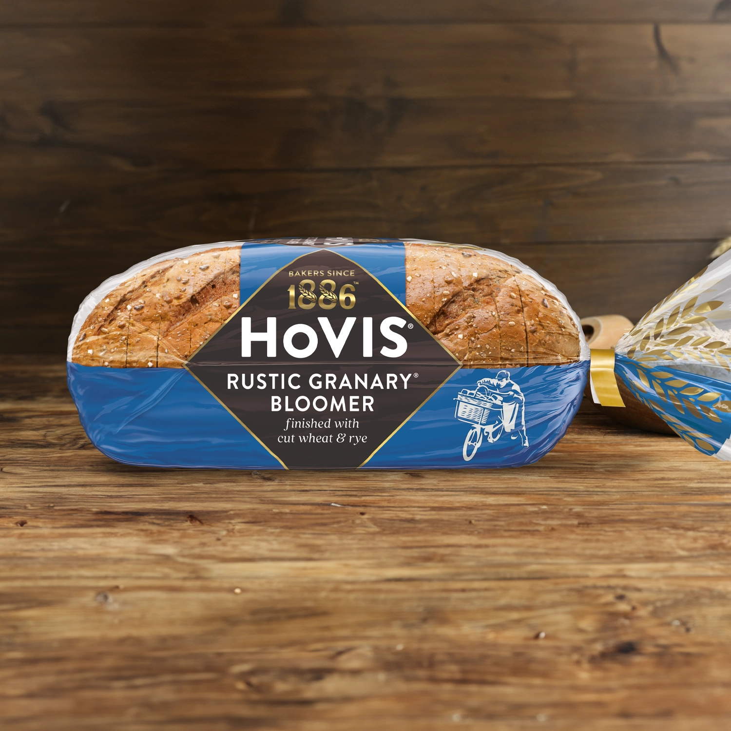 Hovis Granary Bloomer on a wooden table, with a bowl of wheat grains, a wooden scoop, and dried wheat in the background.