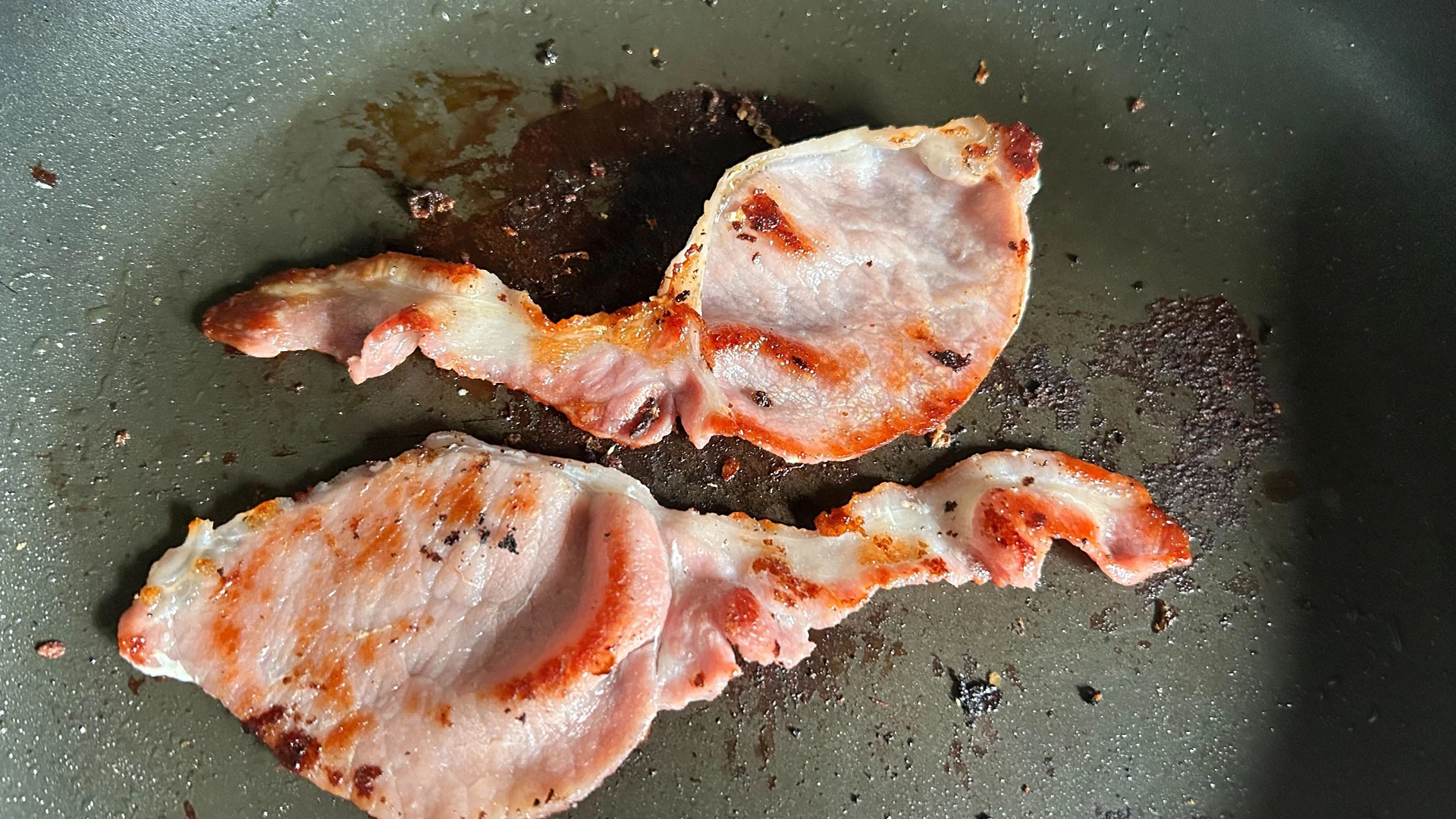 Two slices of bacon, lightly fried, sizzling in a frying pan.