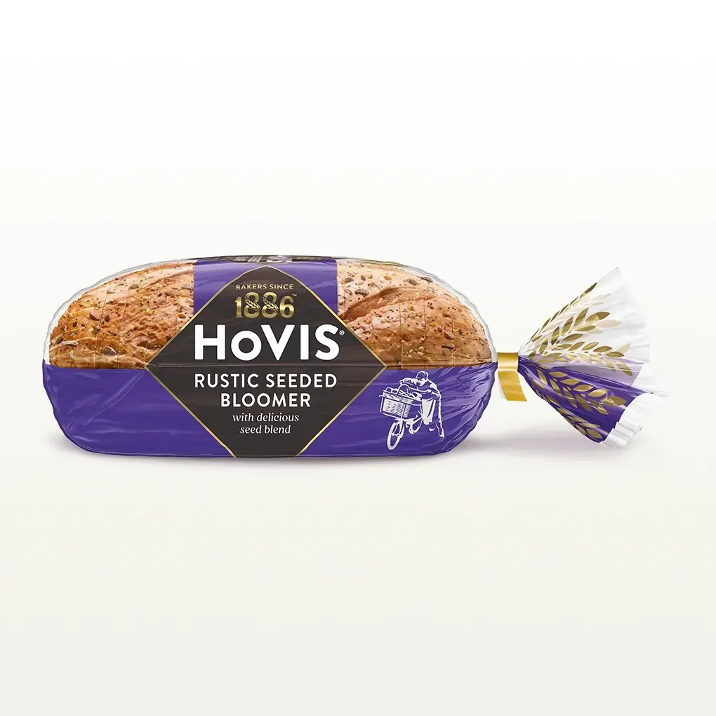 Hovis Seeded Bloomer bread on a white backdrop with a subtle shadow. The packaging is half transparent and half purple, decorated with golden wheats.