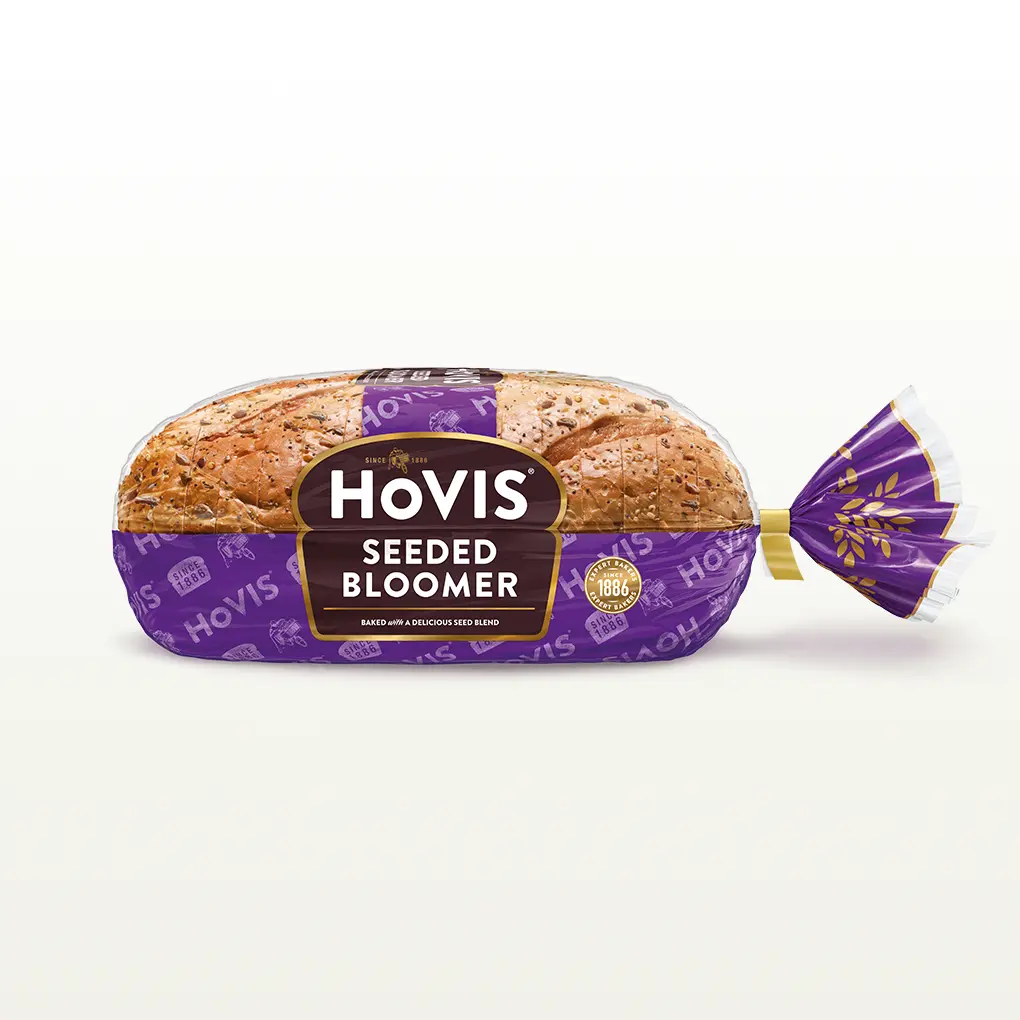 Hovis Seeded Bloomer bread on a white backdrop with a subtle shadow. The packaging is half transparent and half purple, decorated with golden wheats.