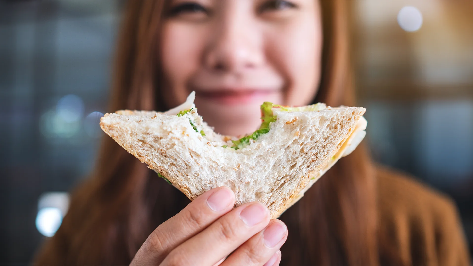 Closeup photo of a woman smiling and holding up a fresh sandwich with a bite out of it.