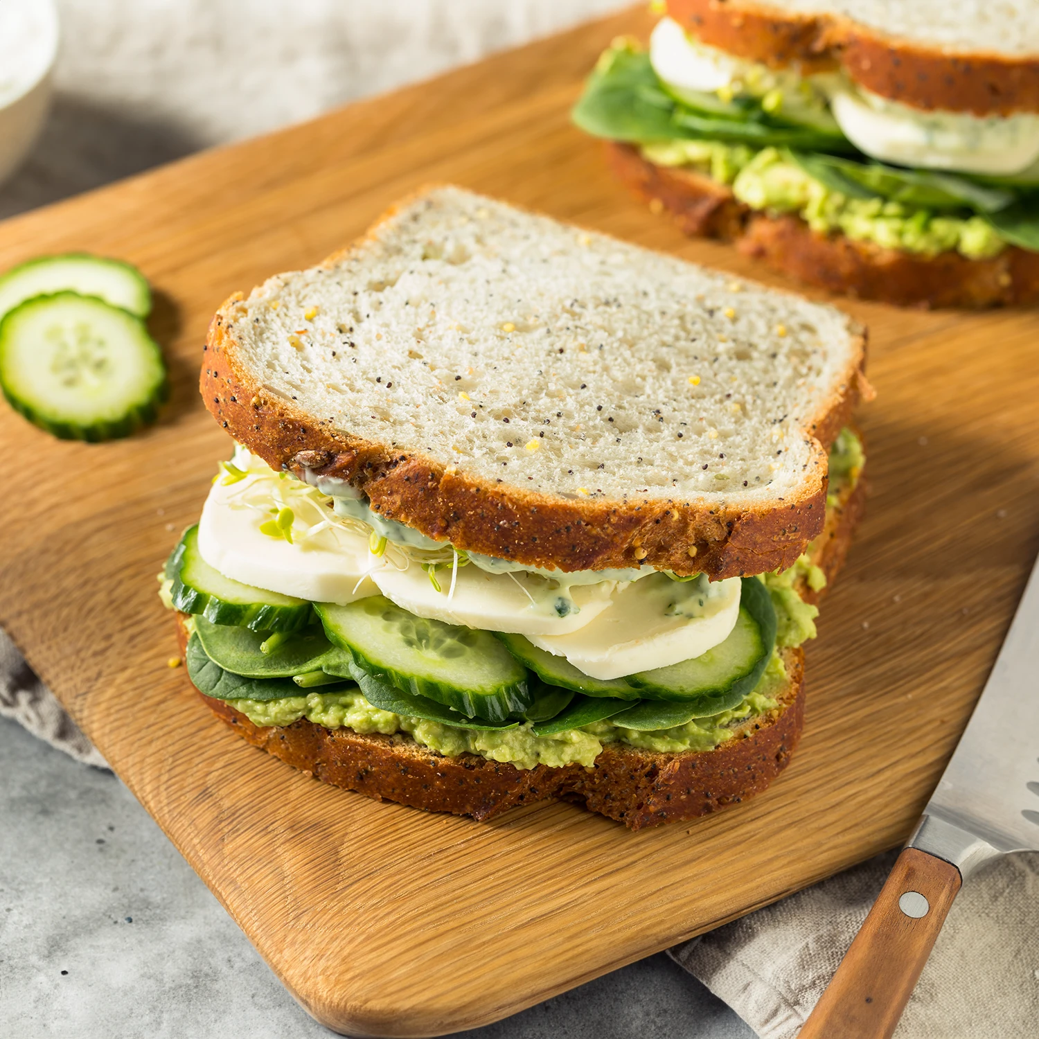 A seeded bread sandwich filled with mashed avocado, sliced cucumber, and mozzarella cheese, with cucumber slices and a knife on the side.