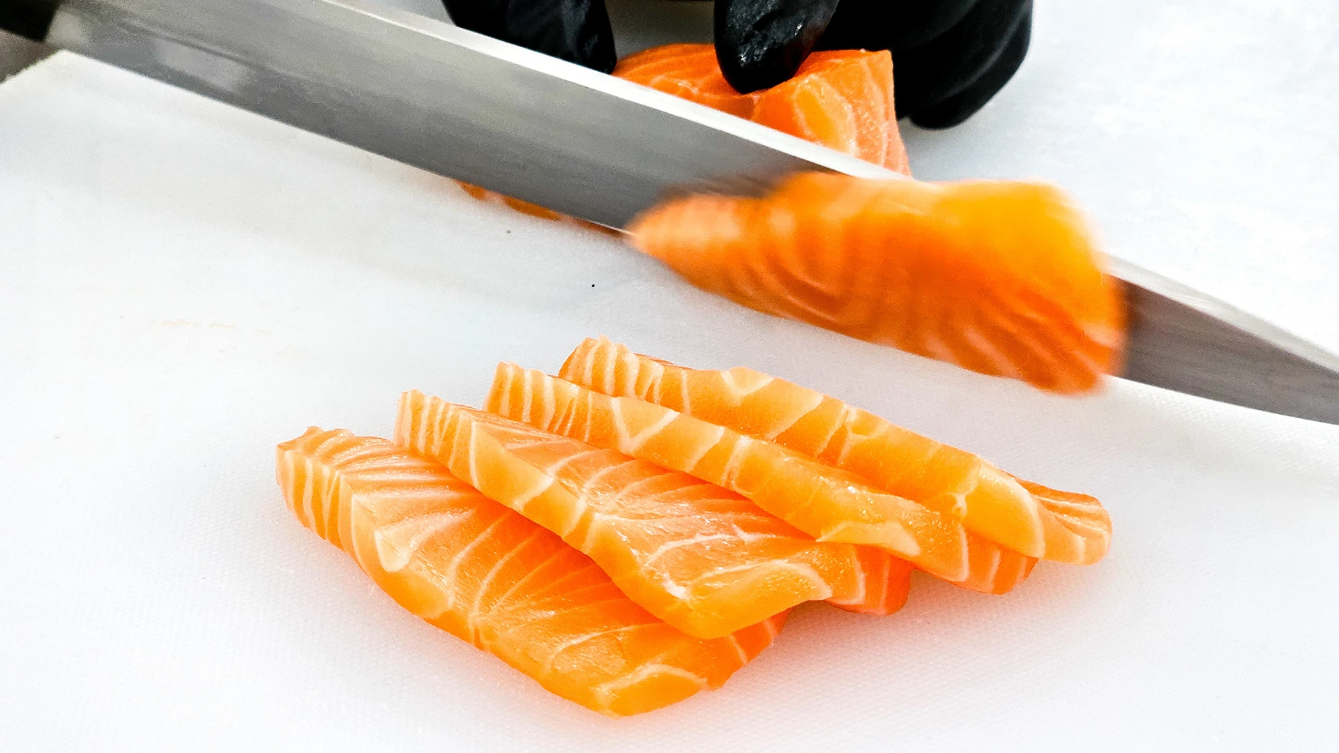 A hand holds a piece of raw salmon on a chopping board while the other hand uses a sharp knife to slice it into thinner pieces.