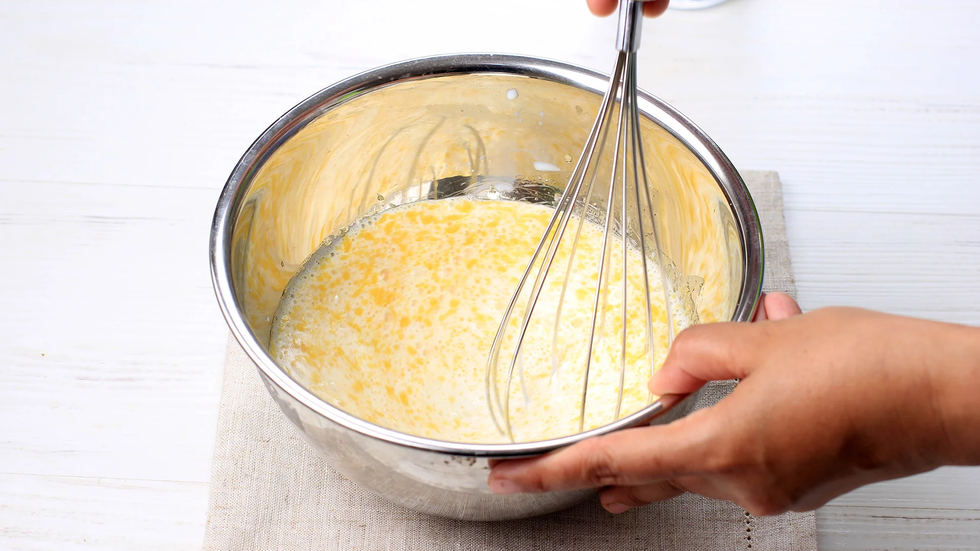 One hand uses a metal whisk to mix eggs, milk, and sugar, while the other holds a medium metal bowl on top of a linen cloth on a white kitchen counter.