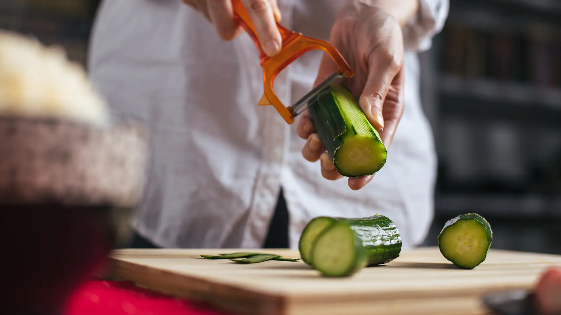 A person using a vegetable peeler to create thin slices of cucumber on a cutting board.