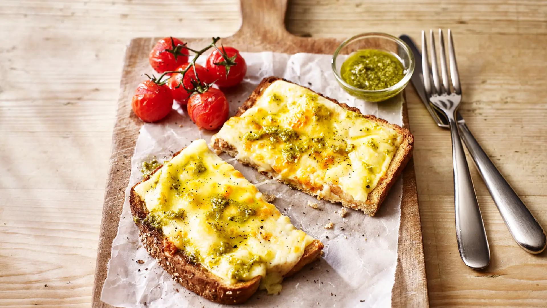 A slice of toasted bread topped with molten cheese and pesto, cut in half, accompanied by grilled cherry tomatoes and a small glass bowl of pesto on the side.