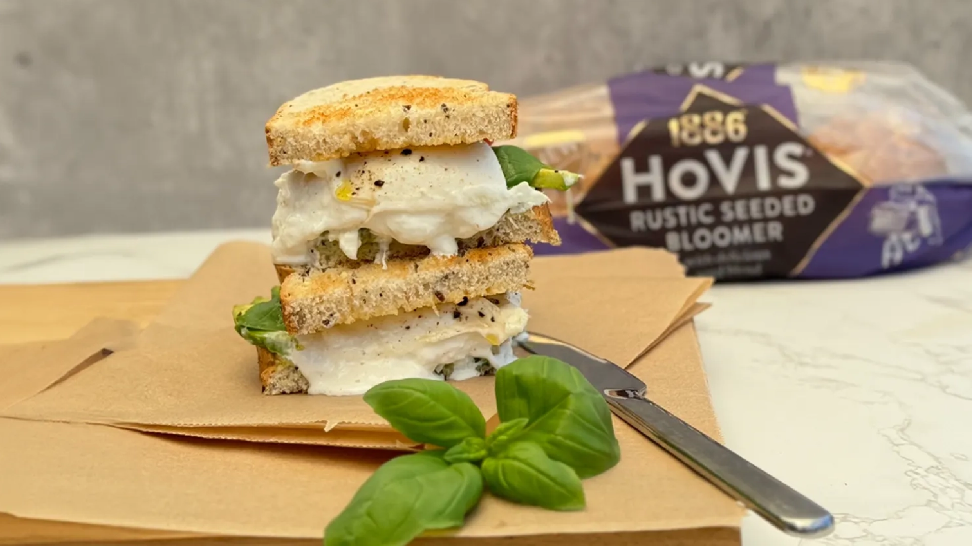 Two halves of a toasted sandwich with creamy white cheese, basil leaves, avocado and small pieces of ground black pepper oozing out. In the foreground, additional basil leaves and a knife are visible.