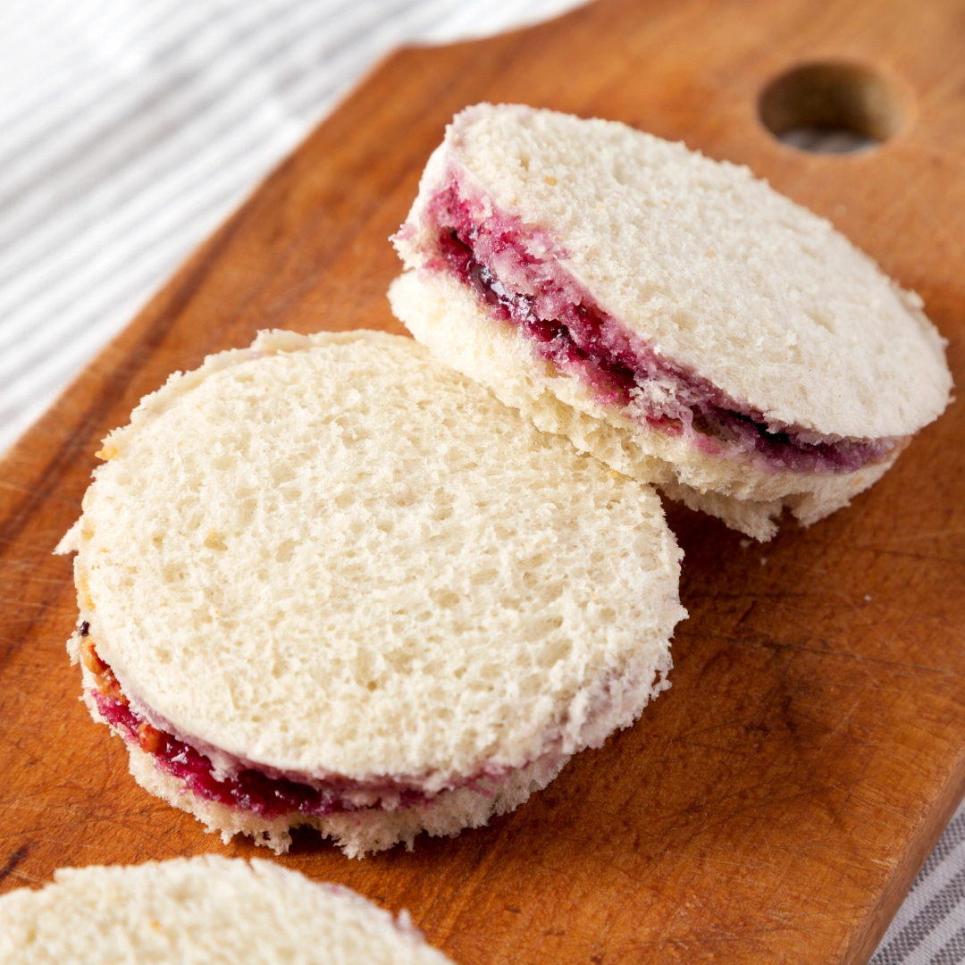 Two small, circular jam sandwiches placed on a wooden chopping board, with the jam slightly visible between the slices of bread.