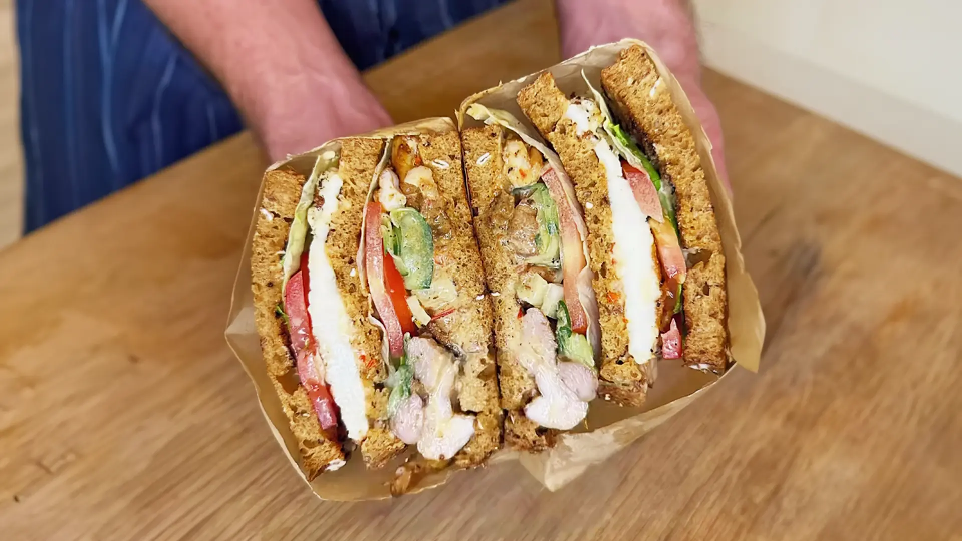 Someone holding two halves of a sandwich wrapped in paper. The sandwich is made of brown bread, filled with tomato slices, lettuce, juicy grilled chicken, and thin cucumber slices.