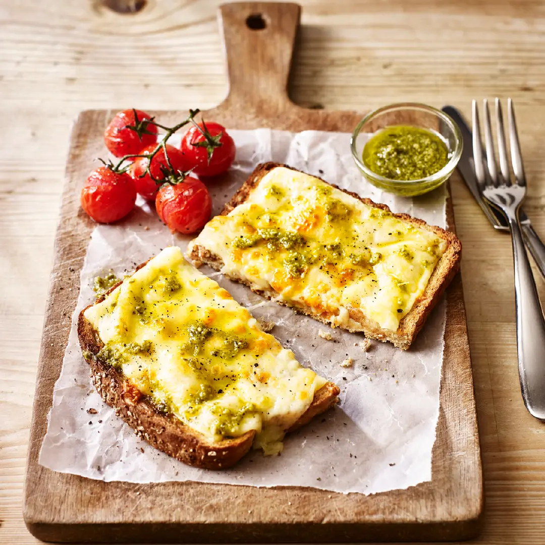 A slice of toasted bread topped with molten cheese and pesto, cut in half, accompanied by grilled cherry tomatoes and a small glass bowl of pesto on the side.