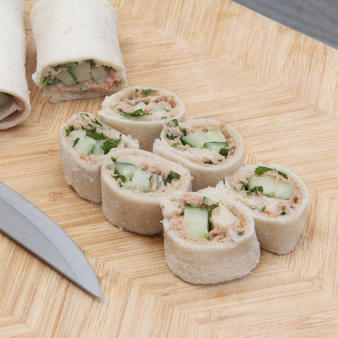 Thin bread wrapped around sliced cucumber and tuna mayo, cut into six bite-sized pieces resembling sushi, with two uncut rolls in the background and a sharp knife.