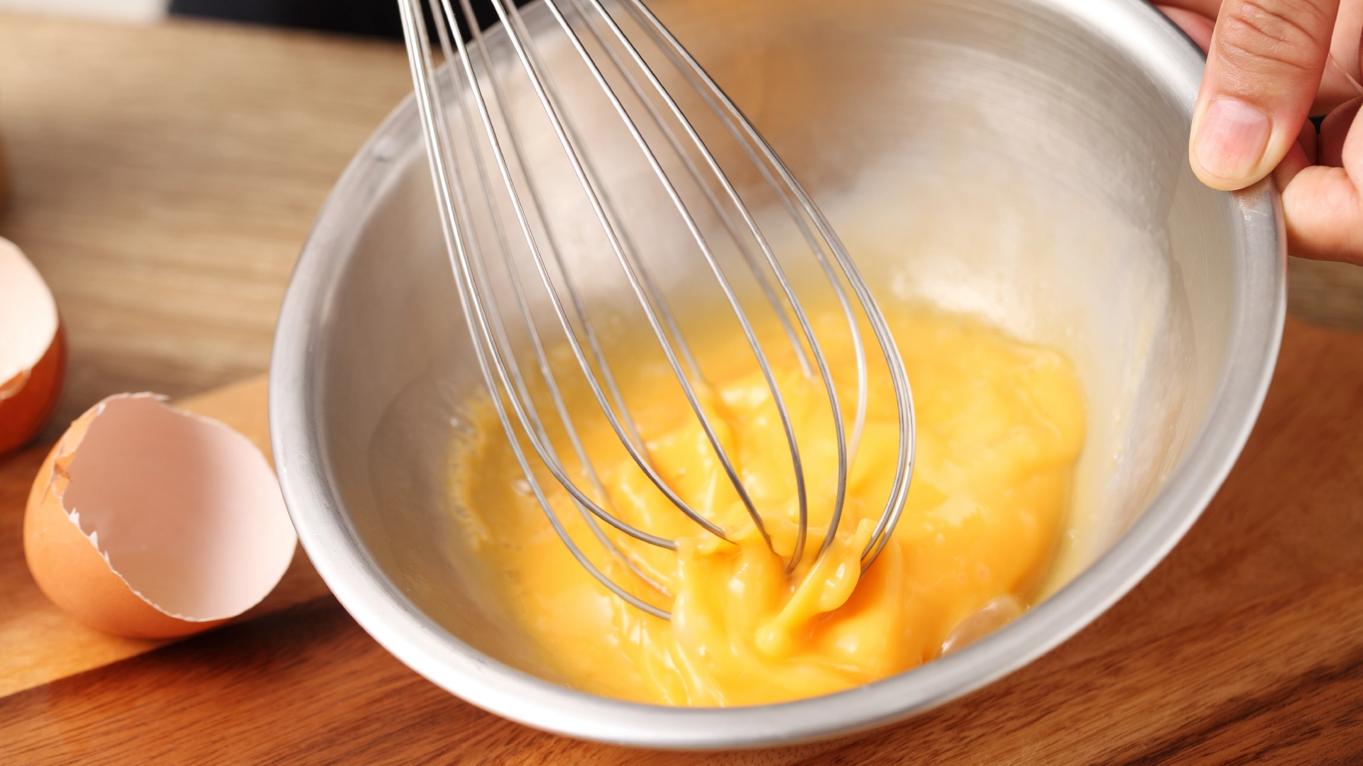 One hand grips a small metal bowl while the other whisks eggs atop a wooden chopping board, with brown cracked egg shells to the side.