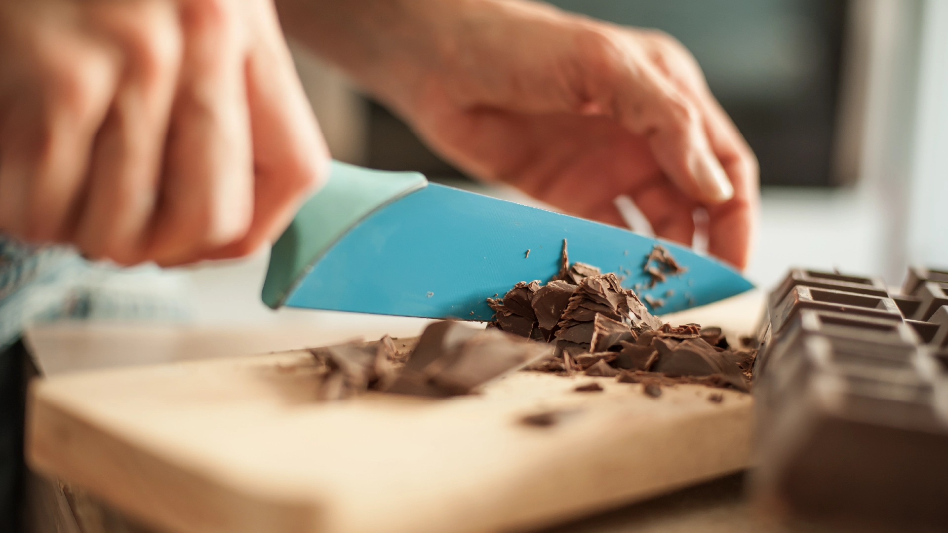 A set of hands chopping a thick chocolate bar on a wooden board.