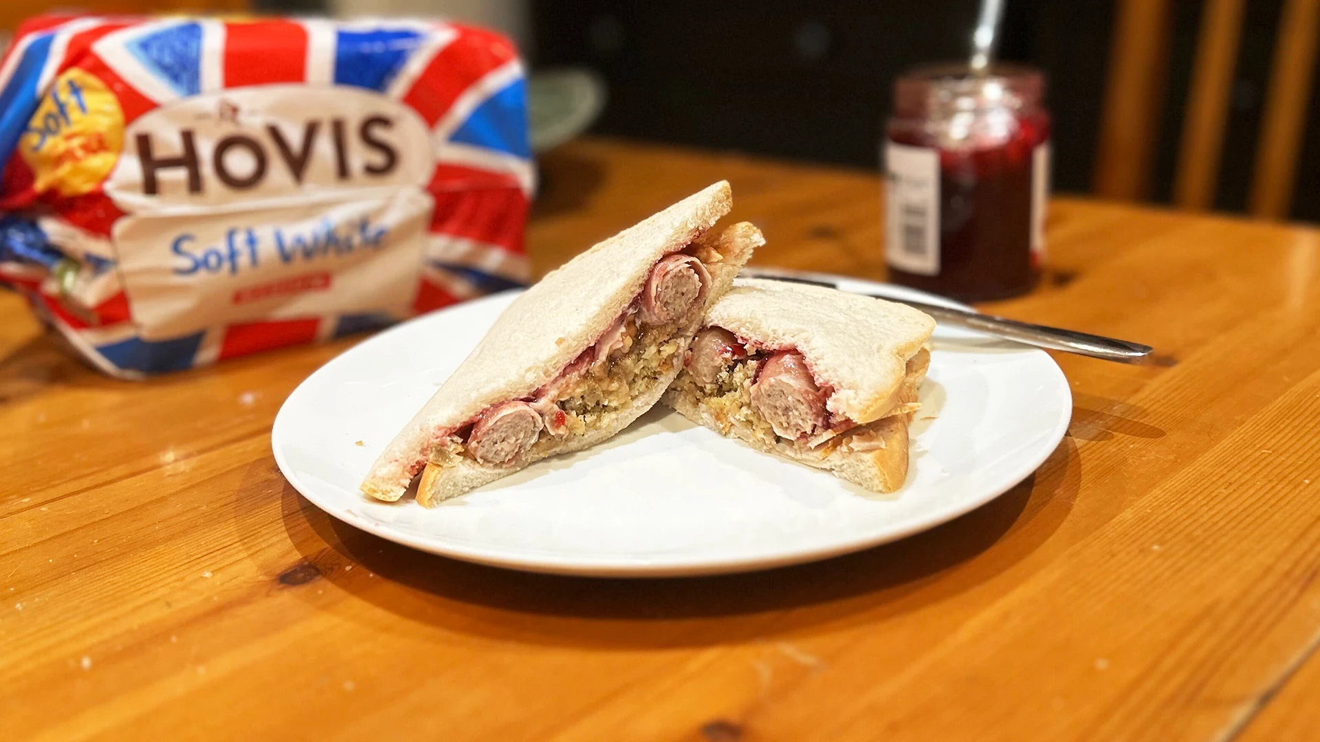 A Hovis Soft White Medium Sliced sandwich sliced in half on a white plate, filled with turkey slices, stuffing, cranberry sauce and pigs in blankets.