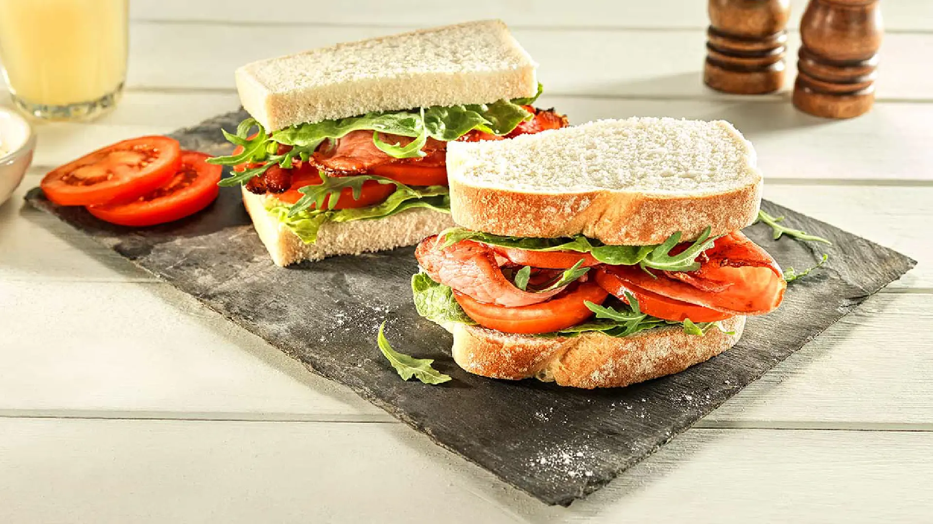 A sandwich cut in half, with crispy bacon, salad leaves and tomato slices between white sliced bread.