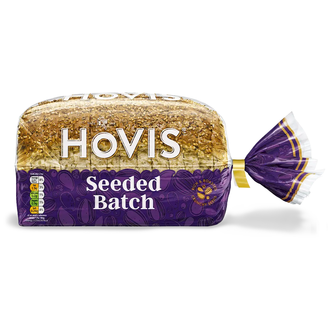 Hovis Seeded Batch bread with a subtle shadow under it. The packaging is half transparent and half purple, featuring images of seeds and a label that reads 'Rich & Roasted, Crunchy Seeds.'