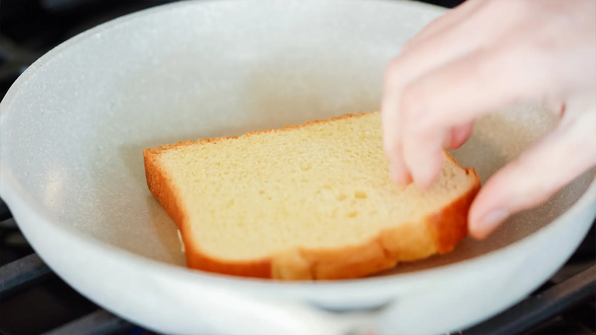 A closed up image of a hand placing a slice of Hovis Farmhouse Batch on a light grey frying pan, on a black hob, under medium heat.