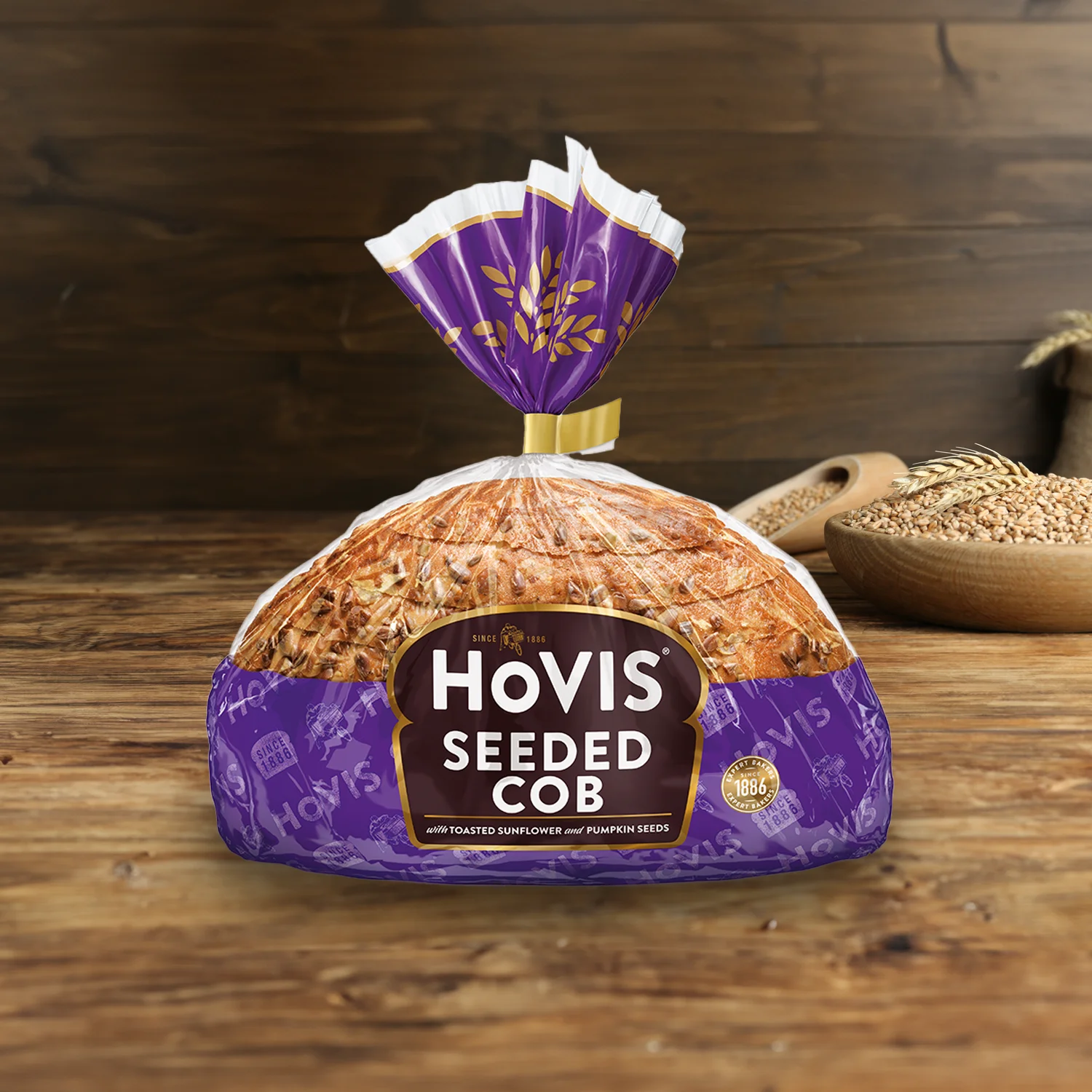 Hovis Seeded Half Cob on a wooden table, with a bowl of wheat grains, a wooden scoop, and dried wheat in the background.