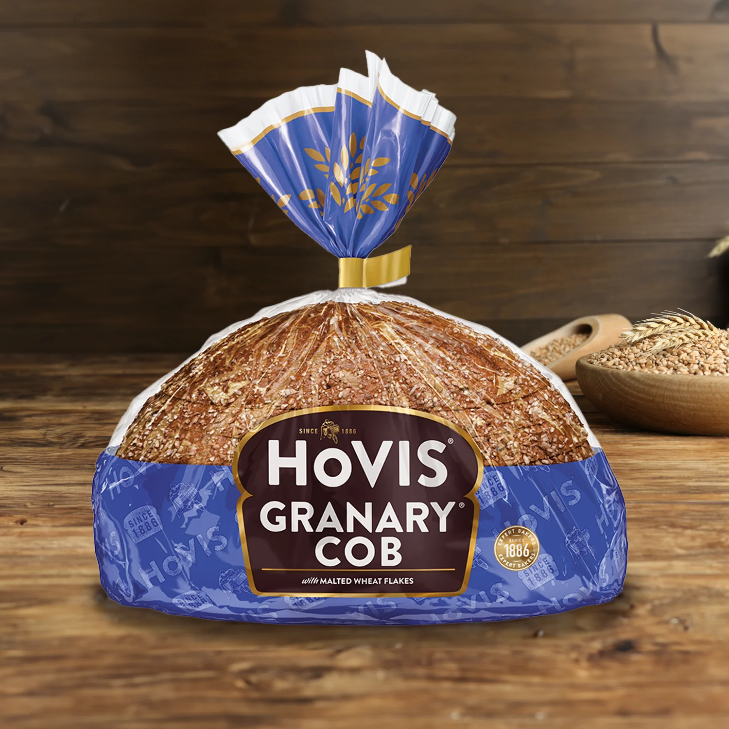 Hovis Granary Half Cob on a wooden table, with a bowl of wheat grains, a wooden scoop, and dried wheat in the background.