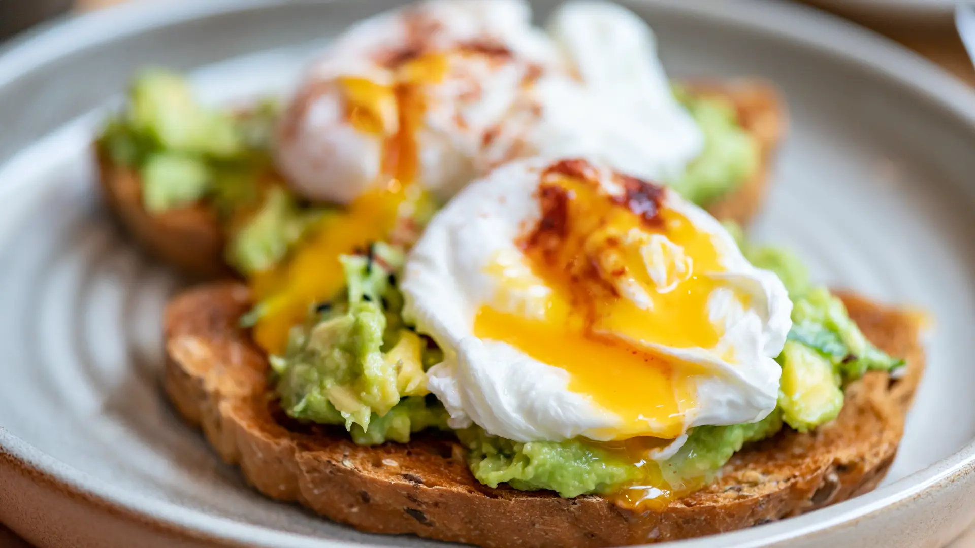 Two slices of slightly toasted seeded bread topped with mashed avocado, a poached egg, and a drizzle of chili oil.