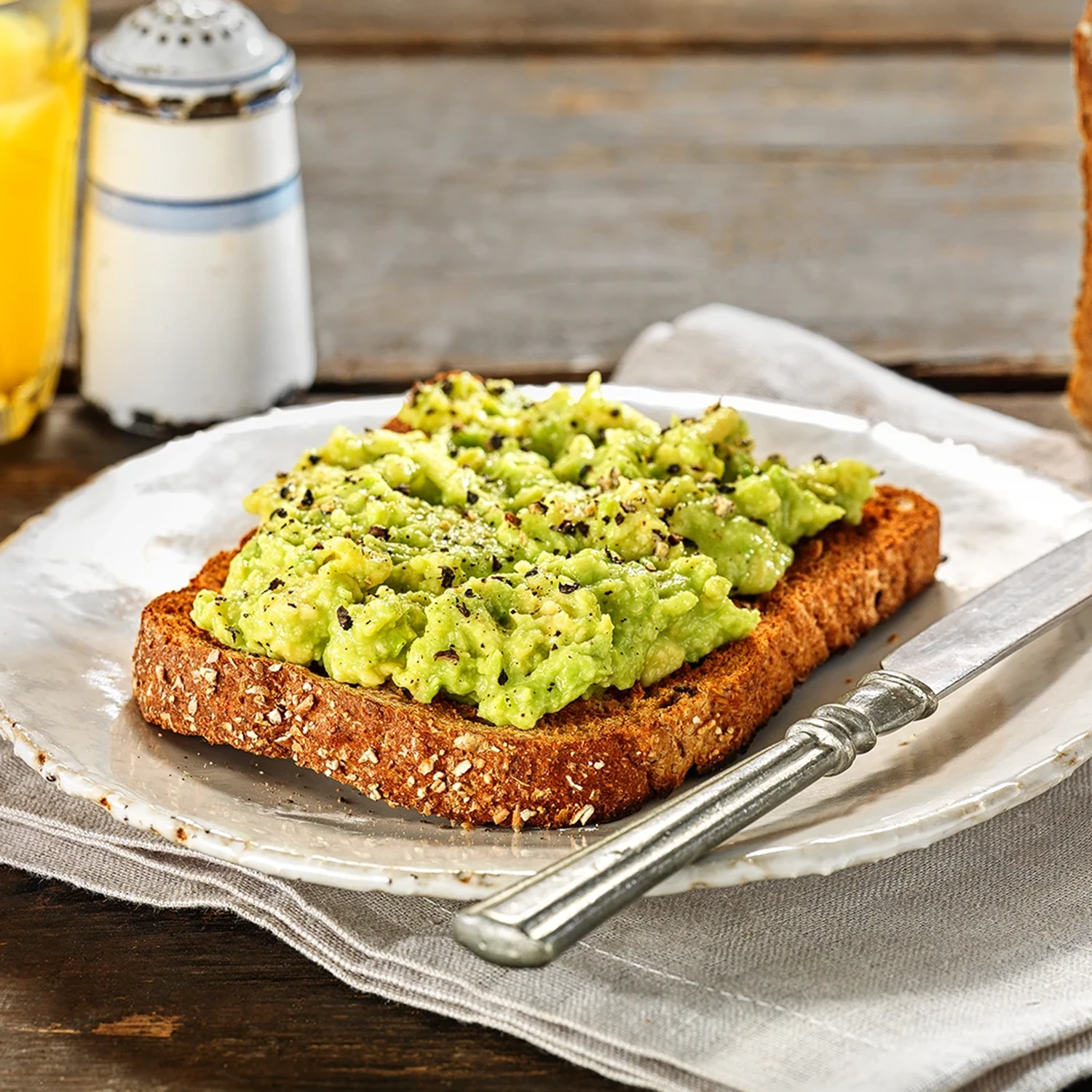 A slice of Hovis Granary Wholemeal bread topped with smashed avocado and a sprinkle of ground black pepper, on a plate with a butter knife on the side.